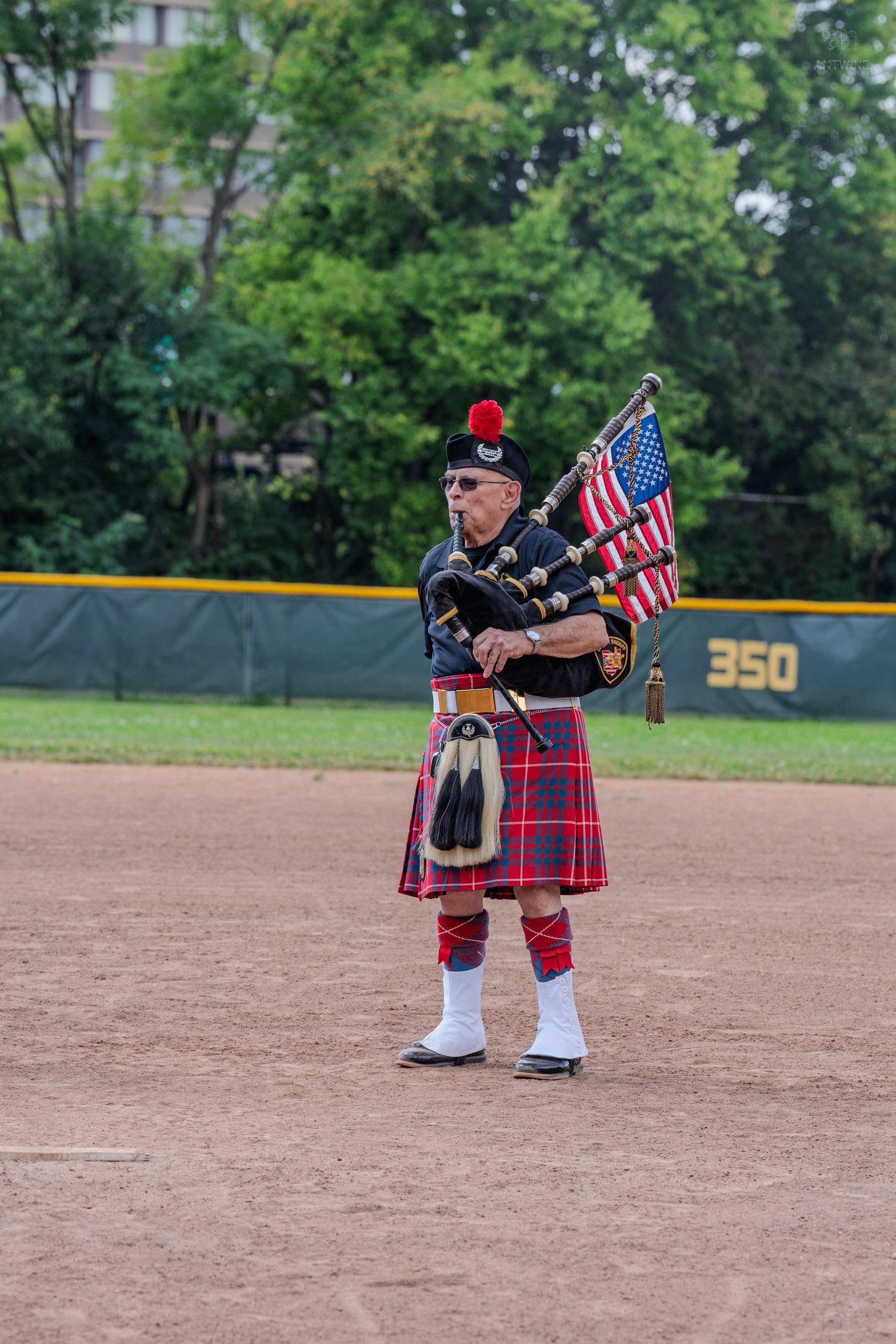 A bagpiper in traditional Scottish tartan kilt and uniform plays bagpipes on a baseball field while holding an American flag.