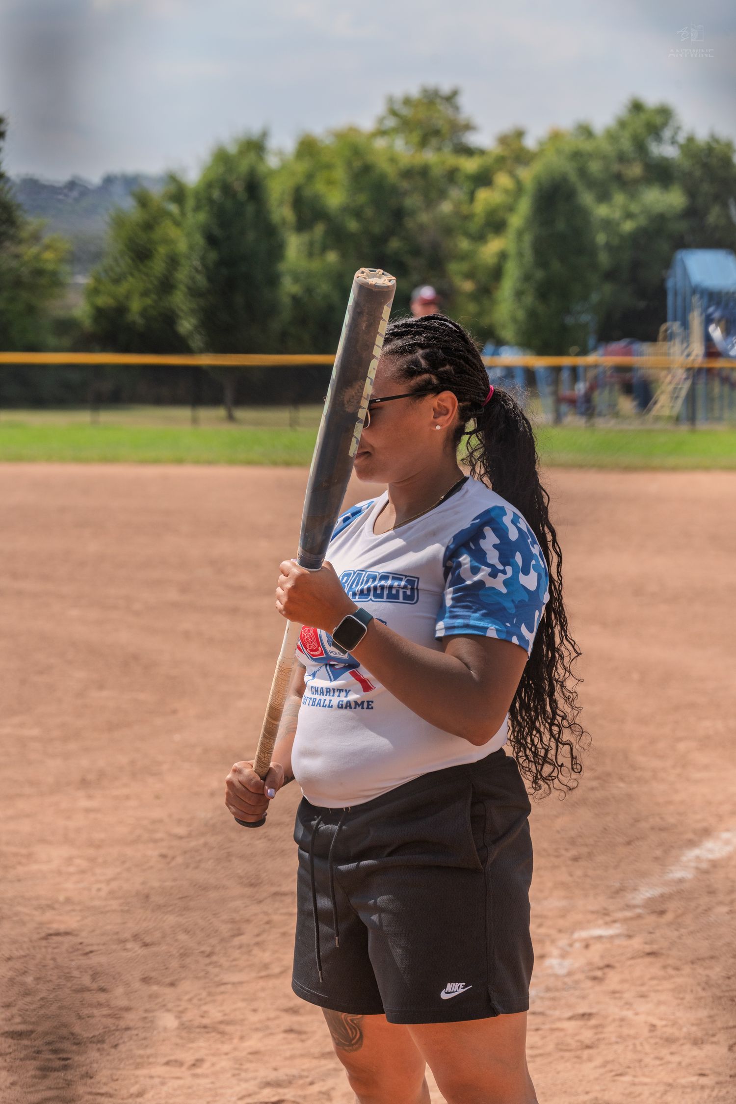Woman with long braids holds a baseball bat ready to bat wearing Nike shorts at an outdoor softball game.