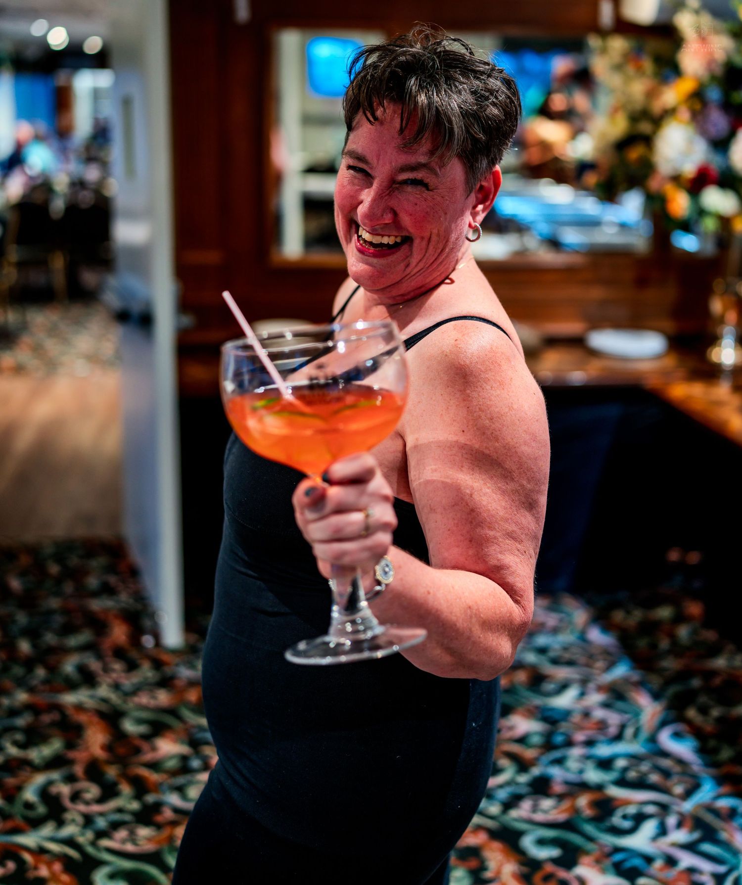 A smiling woman holds a large stemmed glass with an orange cocktail in a restaurant bar setting.