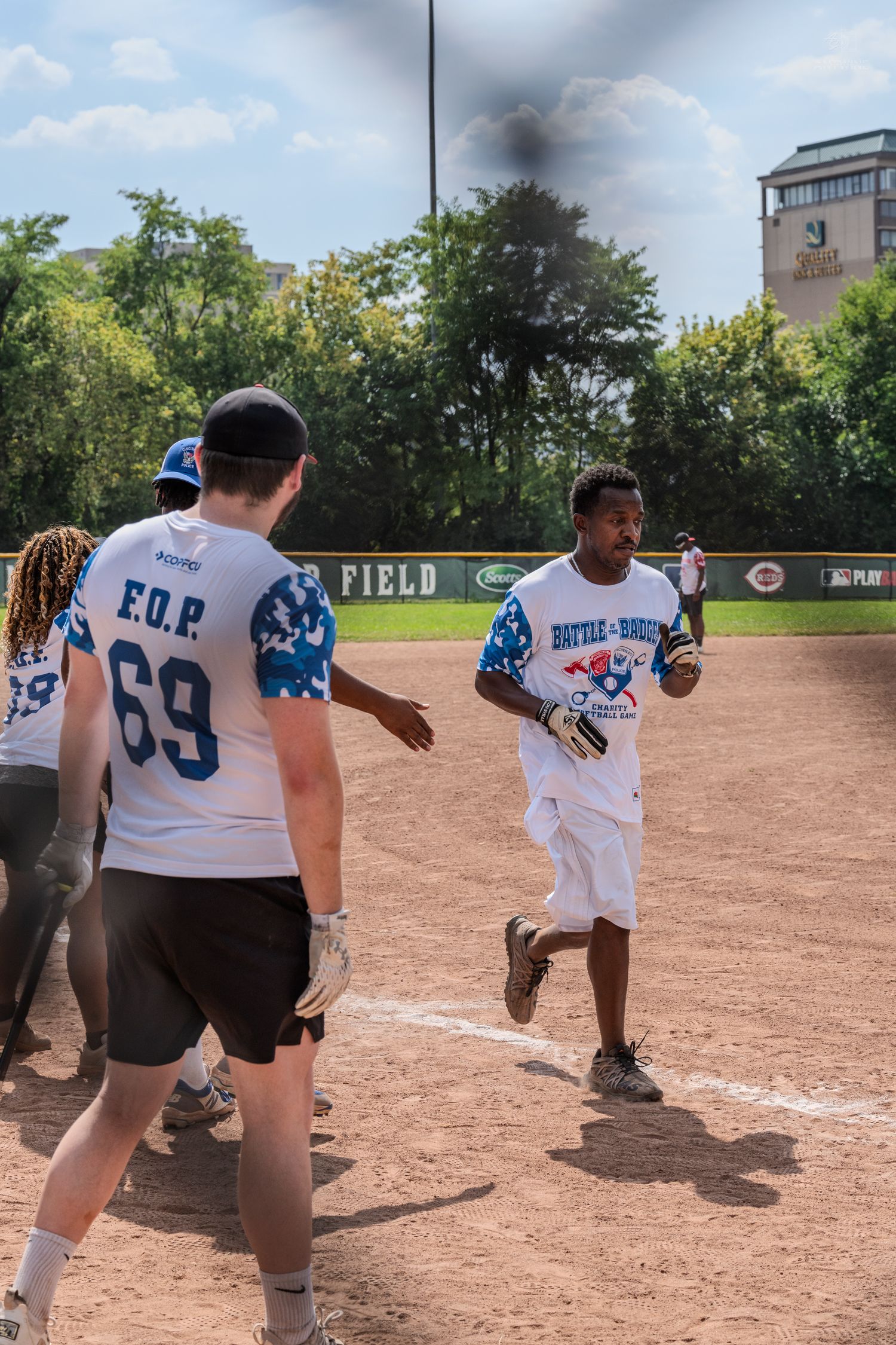 Players celebrate on a baseball diamond during a sunny charity softball game, with player #69 in a blue FOP jersey.