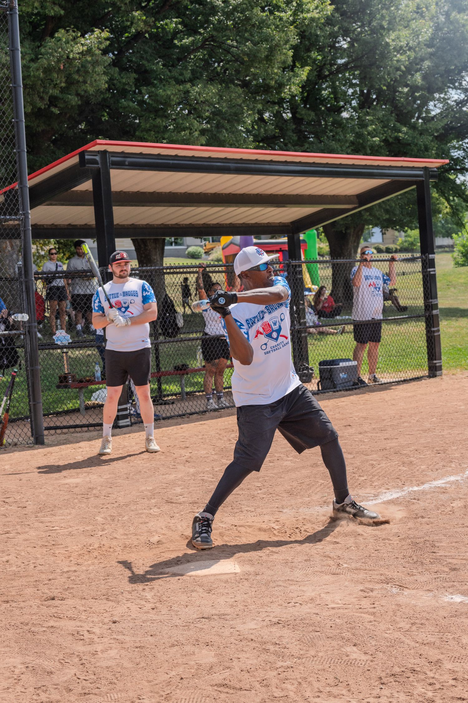 Softball batter swinging at pitch during outdoor charity game with teammates watching from dugout.