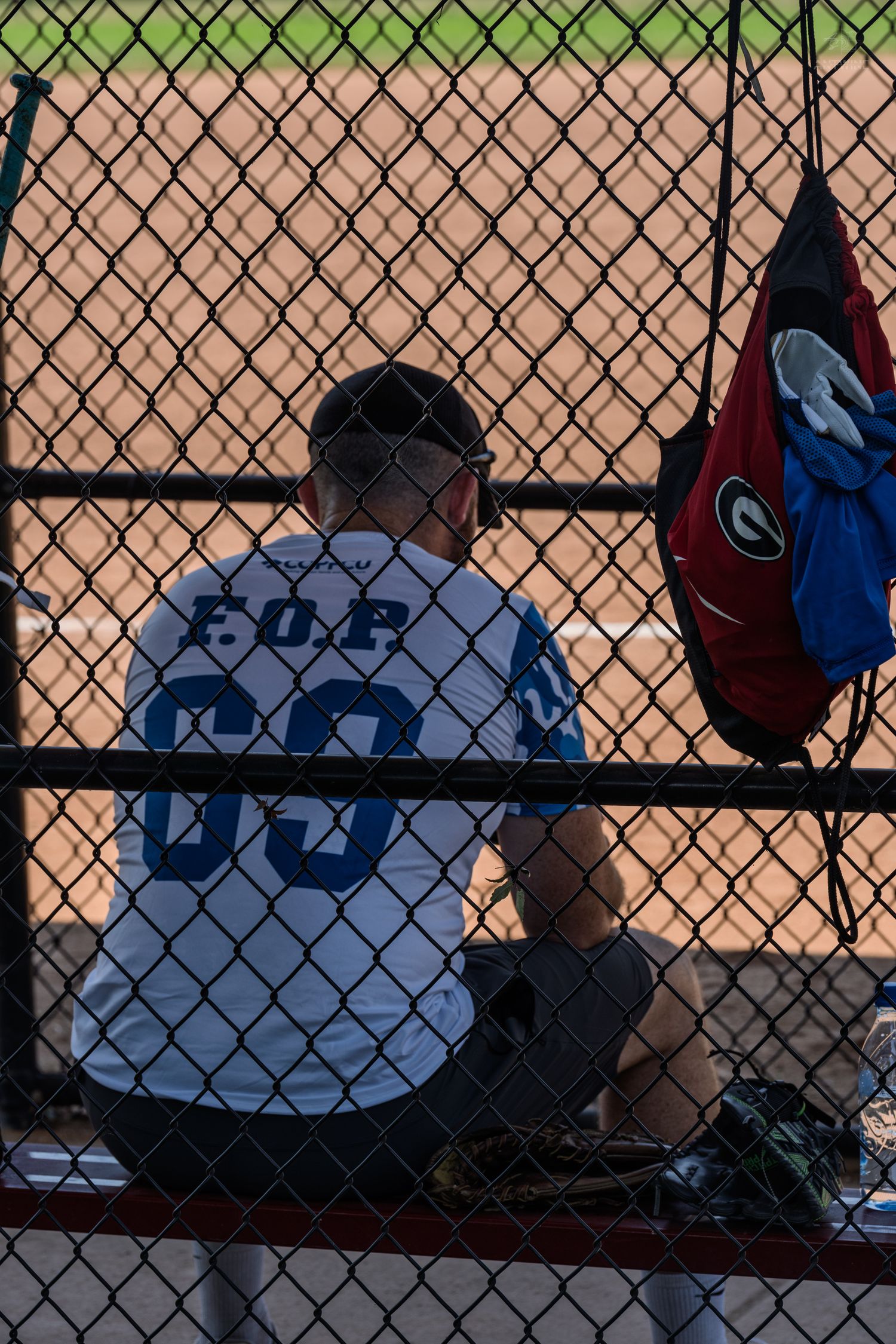 Baseball player in FOP jersey sitting in dugout behind chain-link fence during game.