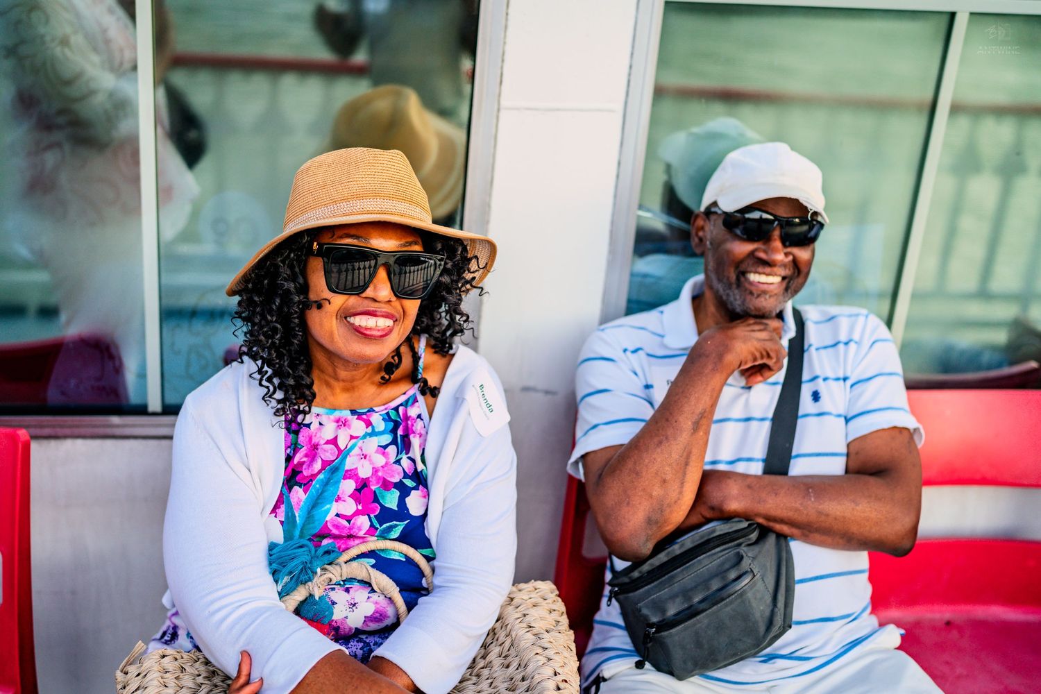 Happy couple wearing sunglasses and hats sitting on red seats outdoors on a sunny day.