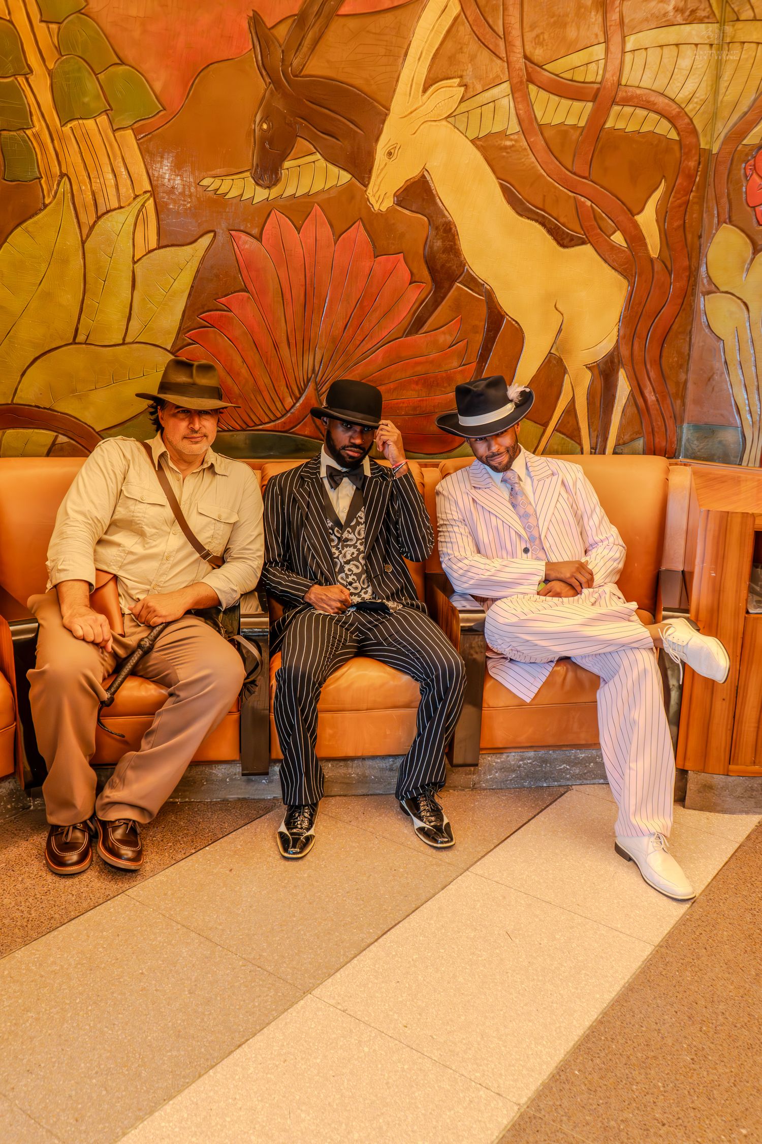 Three stylishly dressed men in vintage suits and fedora hats sitting on a bench before a colorful mural.