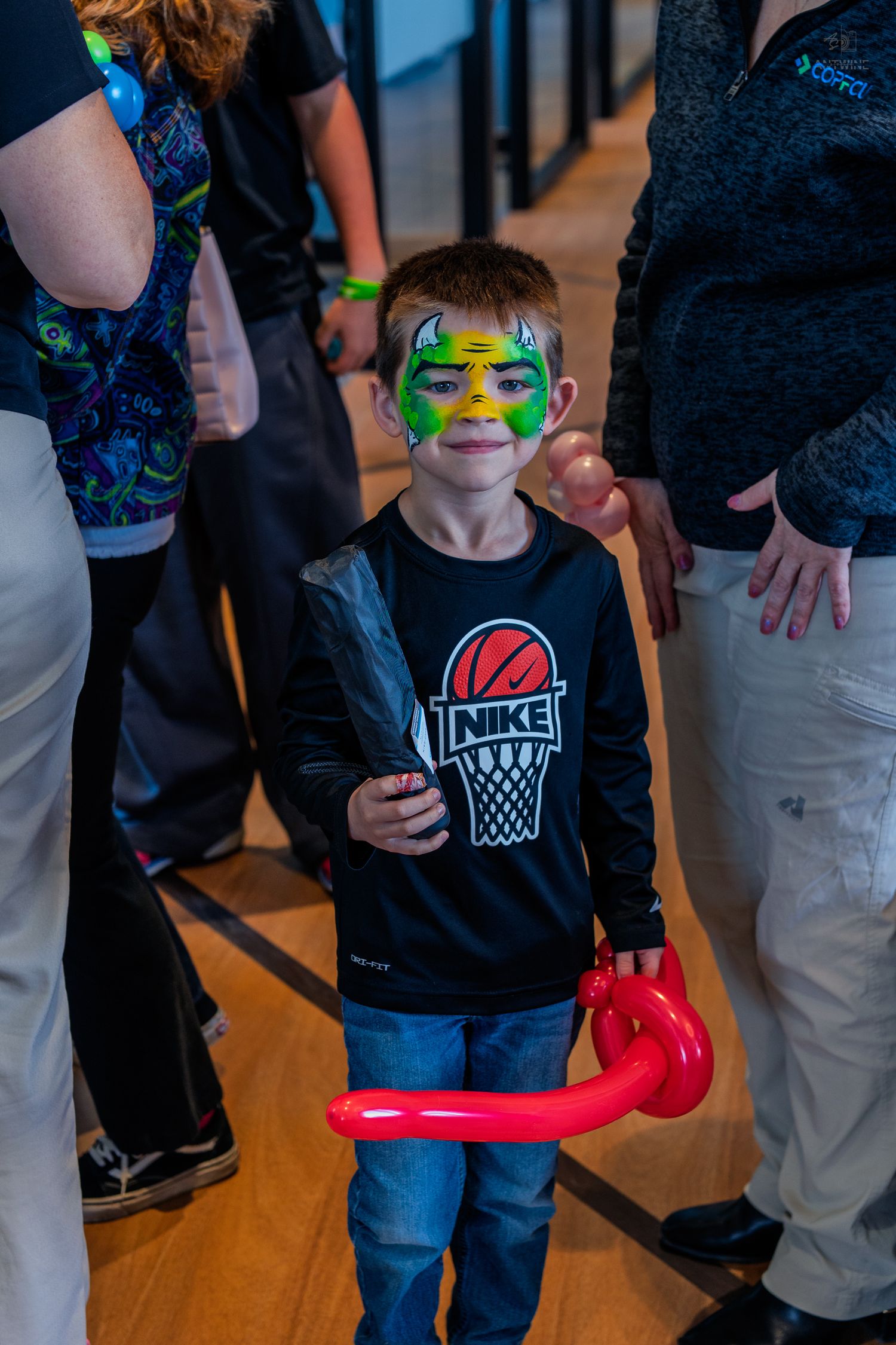 A young boy with green dragon face paint holds a red balloon animal at a festive event, wearing a Nike sweatshirt.