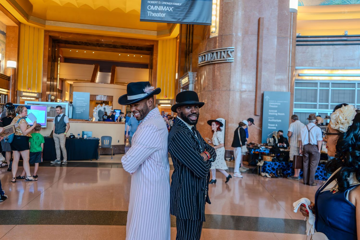 Two men in vintage pinstripe suits and fedora hats pose back-to-back in a grand convention hall lobby.