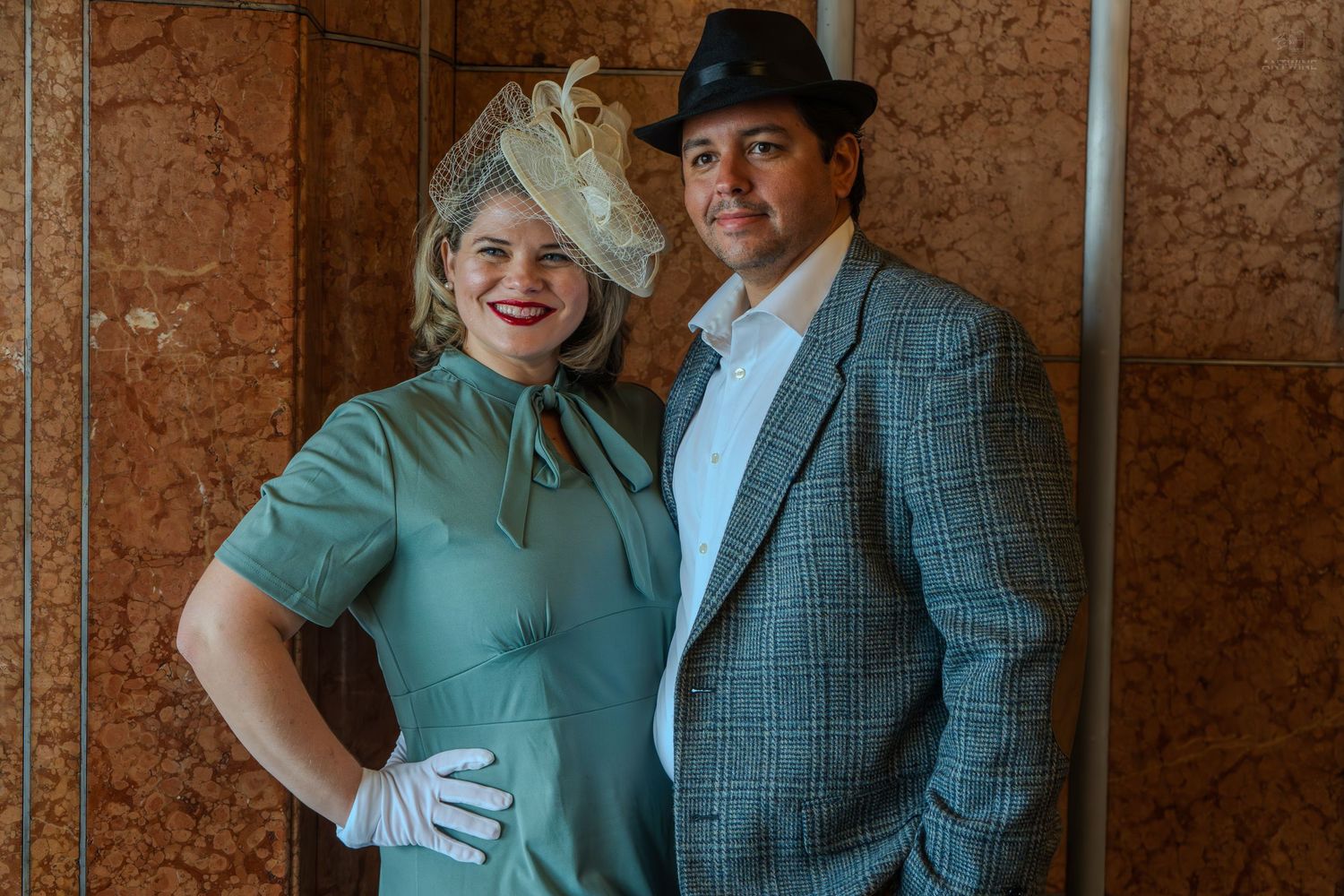 Couple in vintage 1940s attire, woman in green dress with fascinator, man in plaid suit and fedora, posing together.