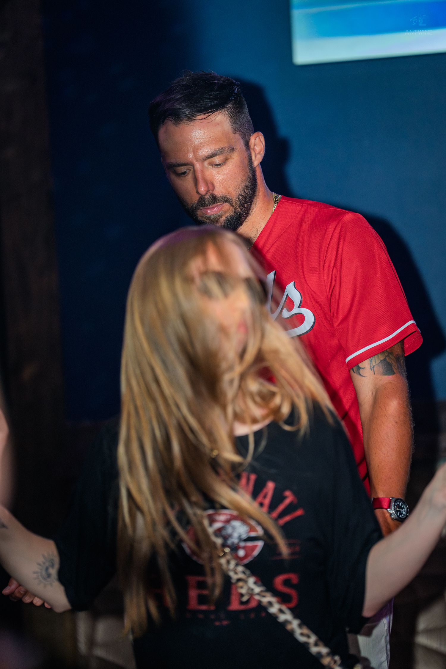 Man in red baseball jersey and woman with blonde hair socializing at a dimly lit indoor event or nightclub.
