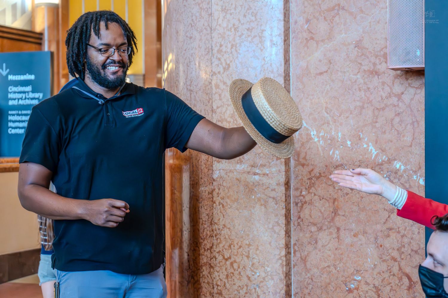 Man in black polo shirt tossing a straw hat to another person inside a marble-walled building.