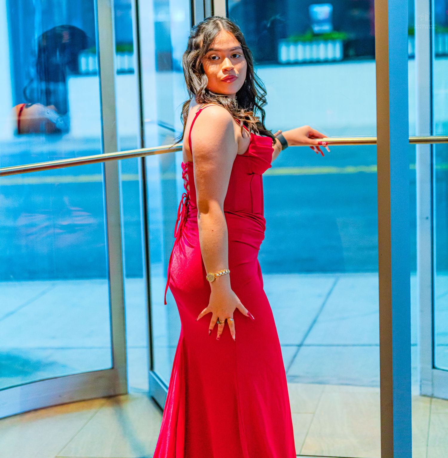 Woman in red mermaid gown poses sideways at glass entrance with gold accessories.
