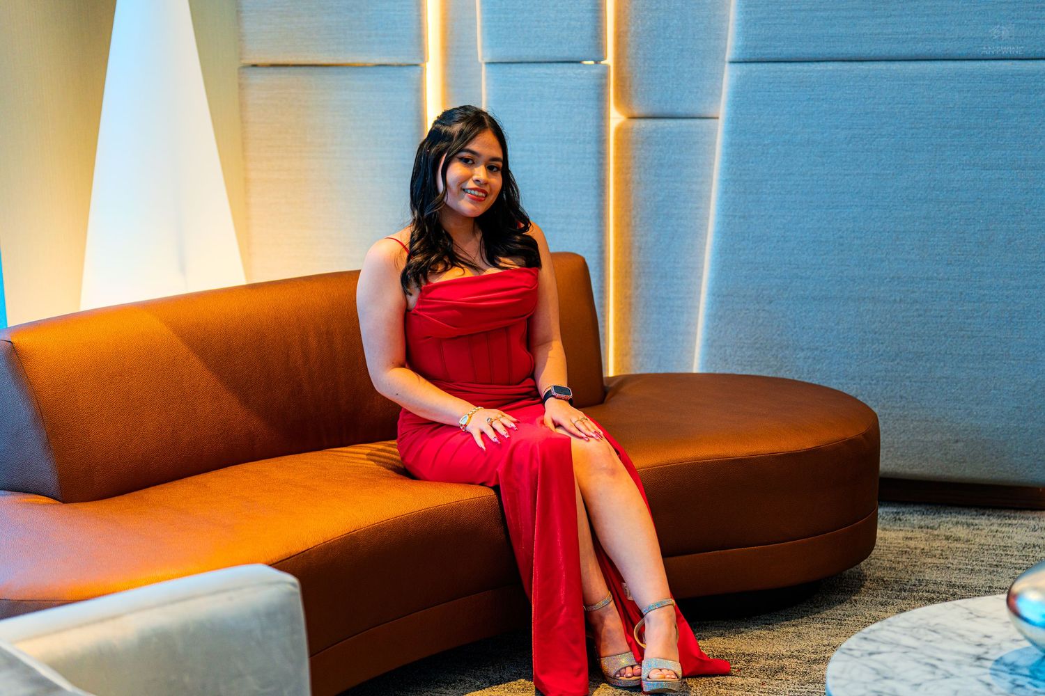 Young woman in a red formal gown sitting gracefully on a curved sofa in an elegant venue.
