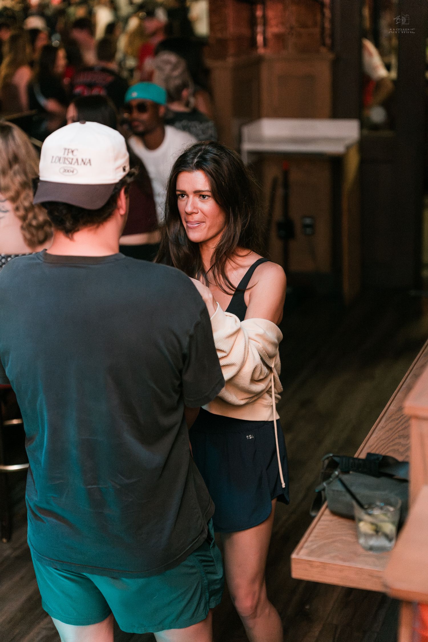 A brunette woman in a black top and shorts converses with a man in a white cap at a busy bar.