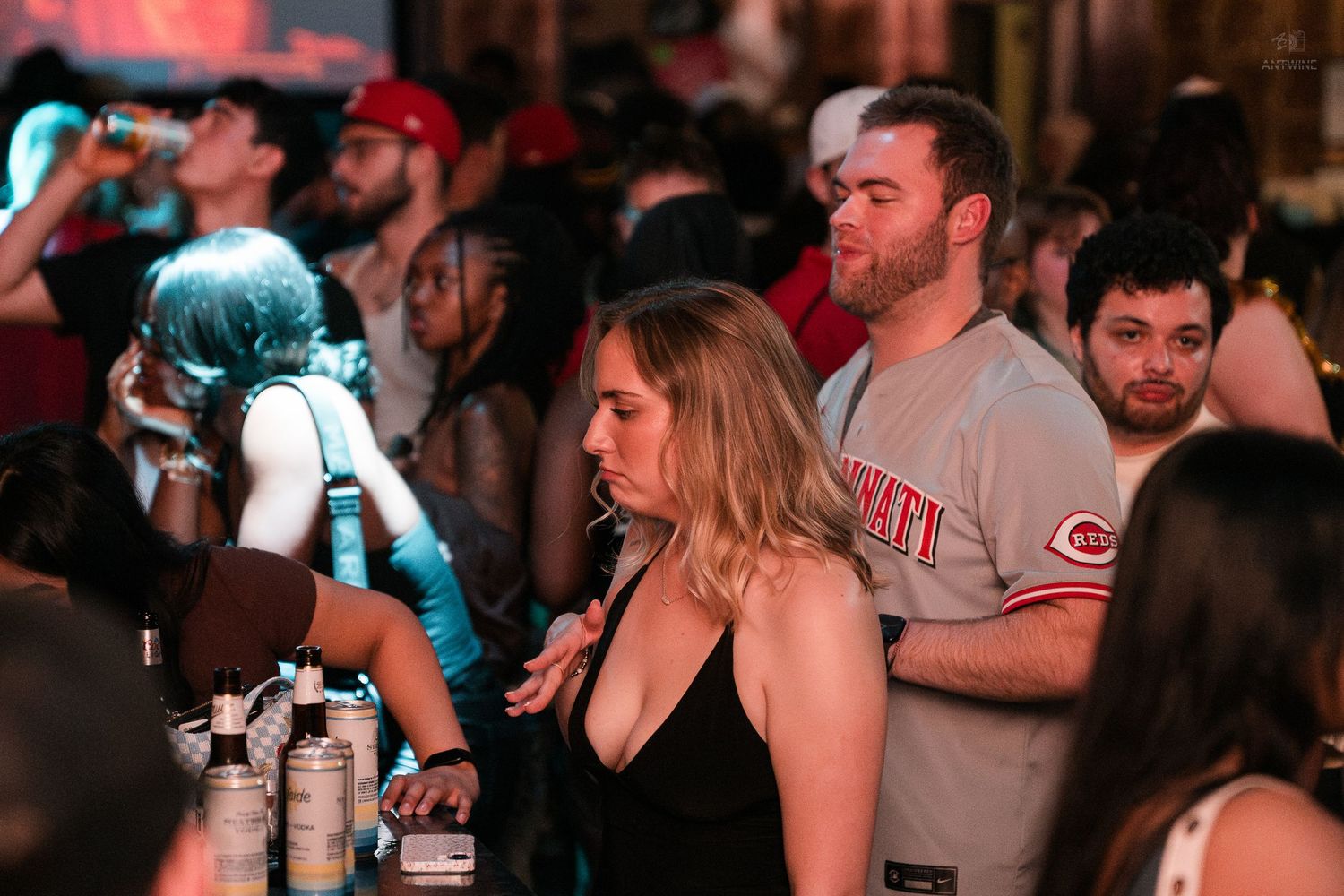 Man in Cincinnati Reds jersey standing at a busy nightclub bar counter.