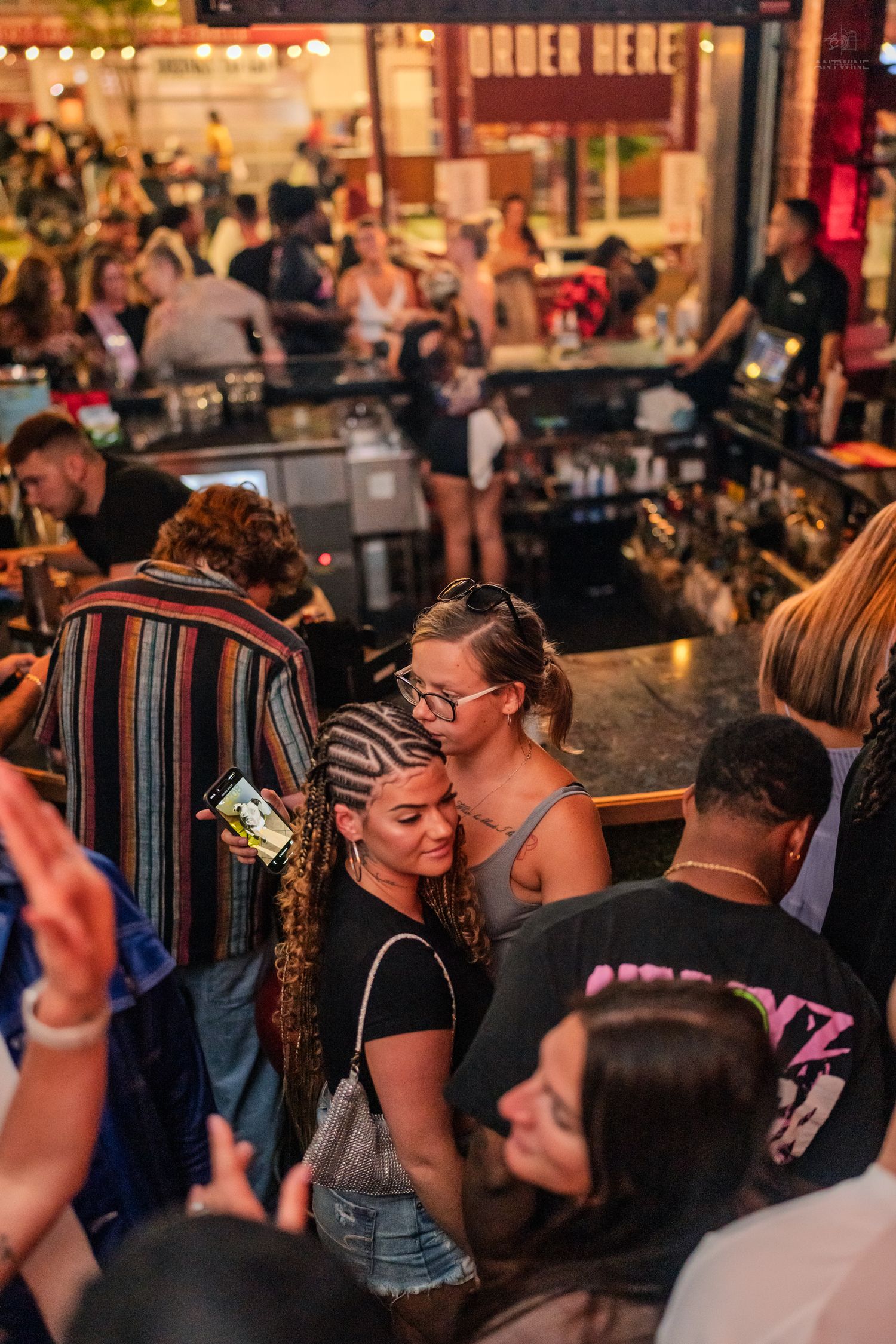 Busy bar crowd with two women taking a selfie near an Order Here sign.
