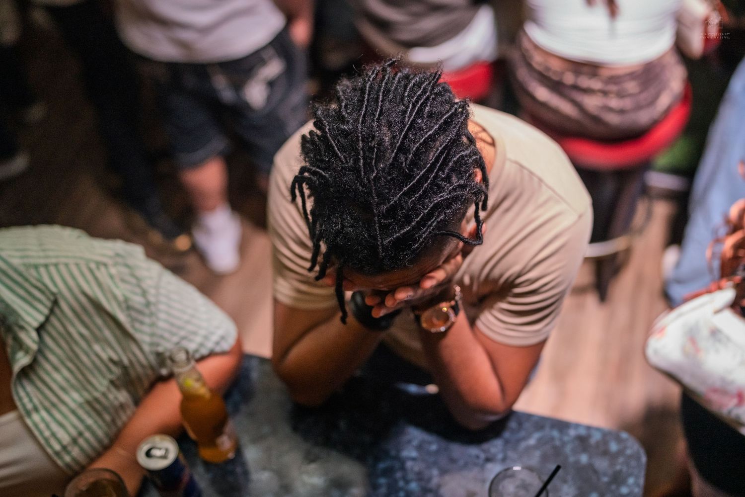 Person with braids resting head on hands at a crowded nightclub, drinks nearby.