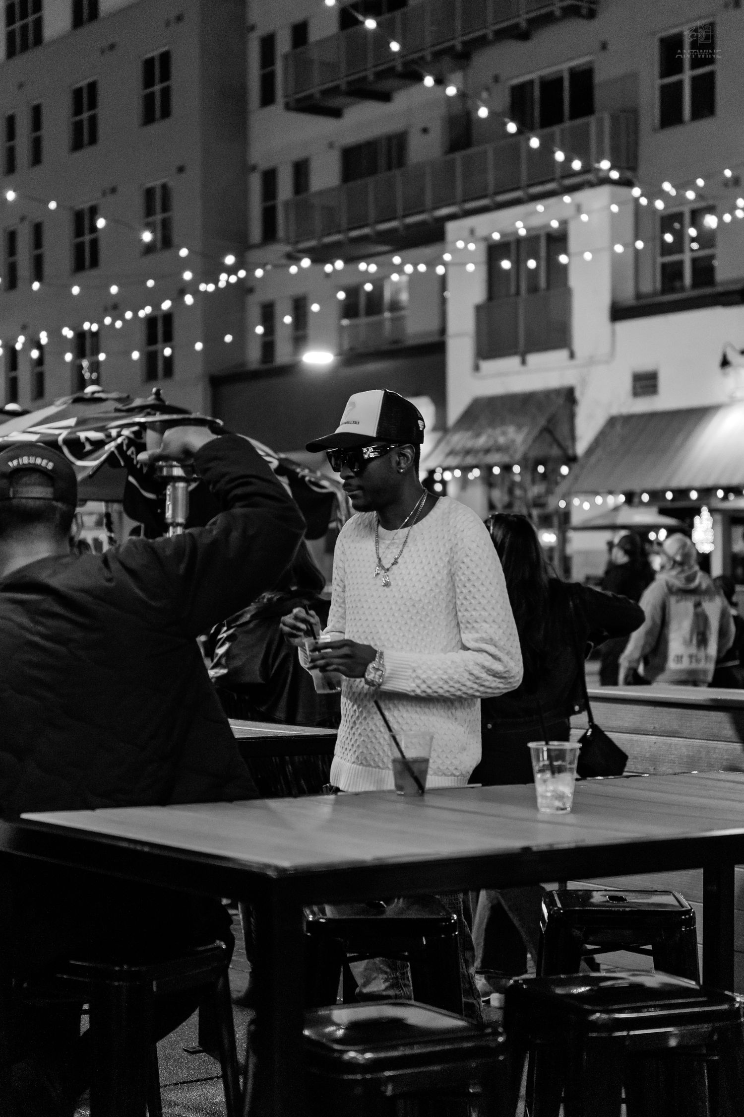 Black and white photo of a man in a white sweater and cap standing at an outdoor bar table.