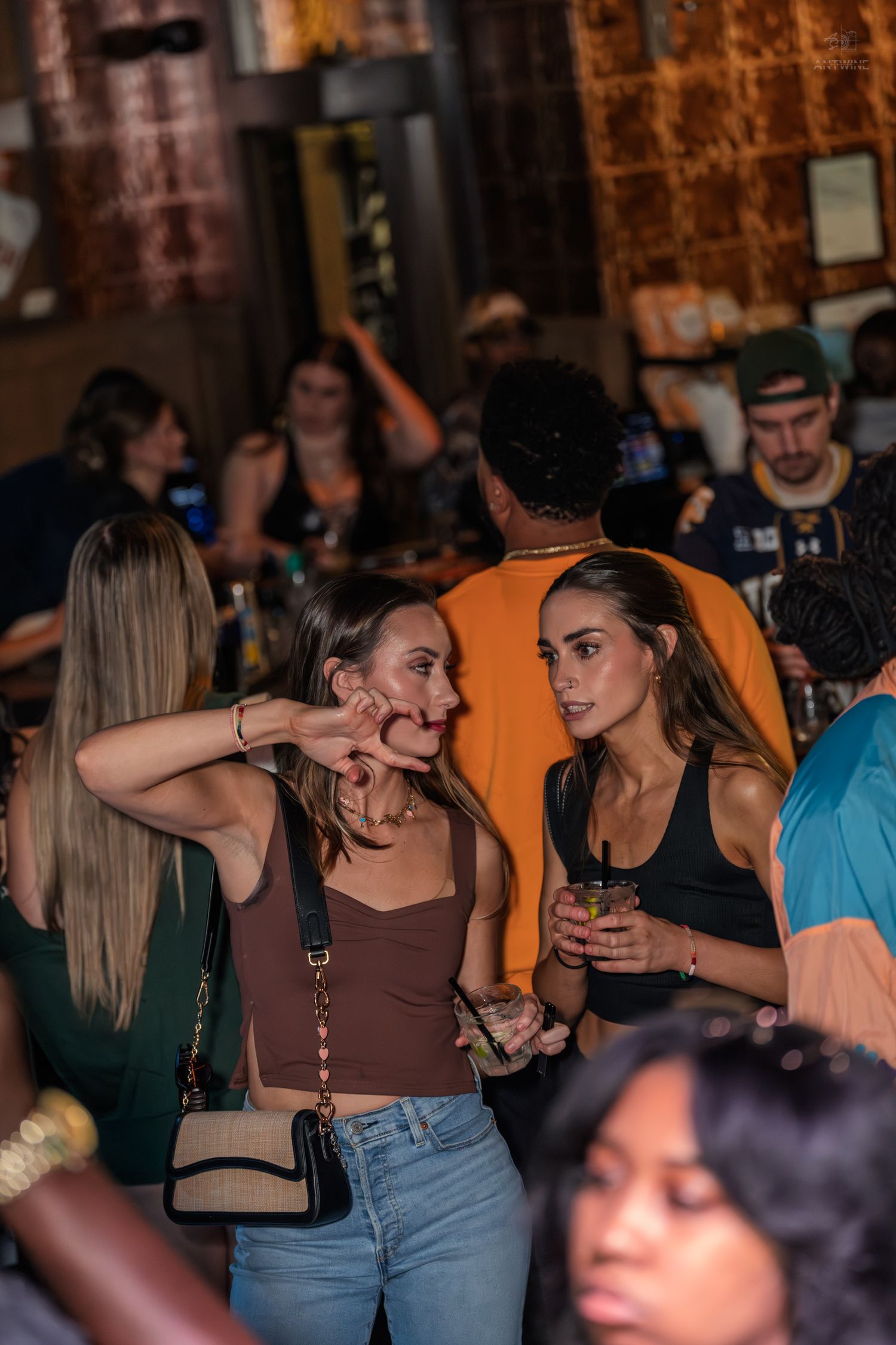 Two women socializing at a crowded bar, one in a brown crop top with crossbody bag, the other in a black tank top holding a drink.
