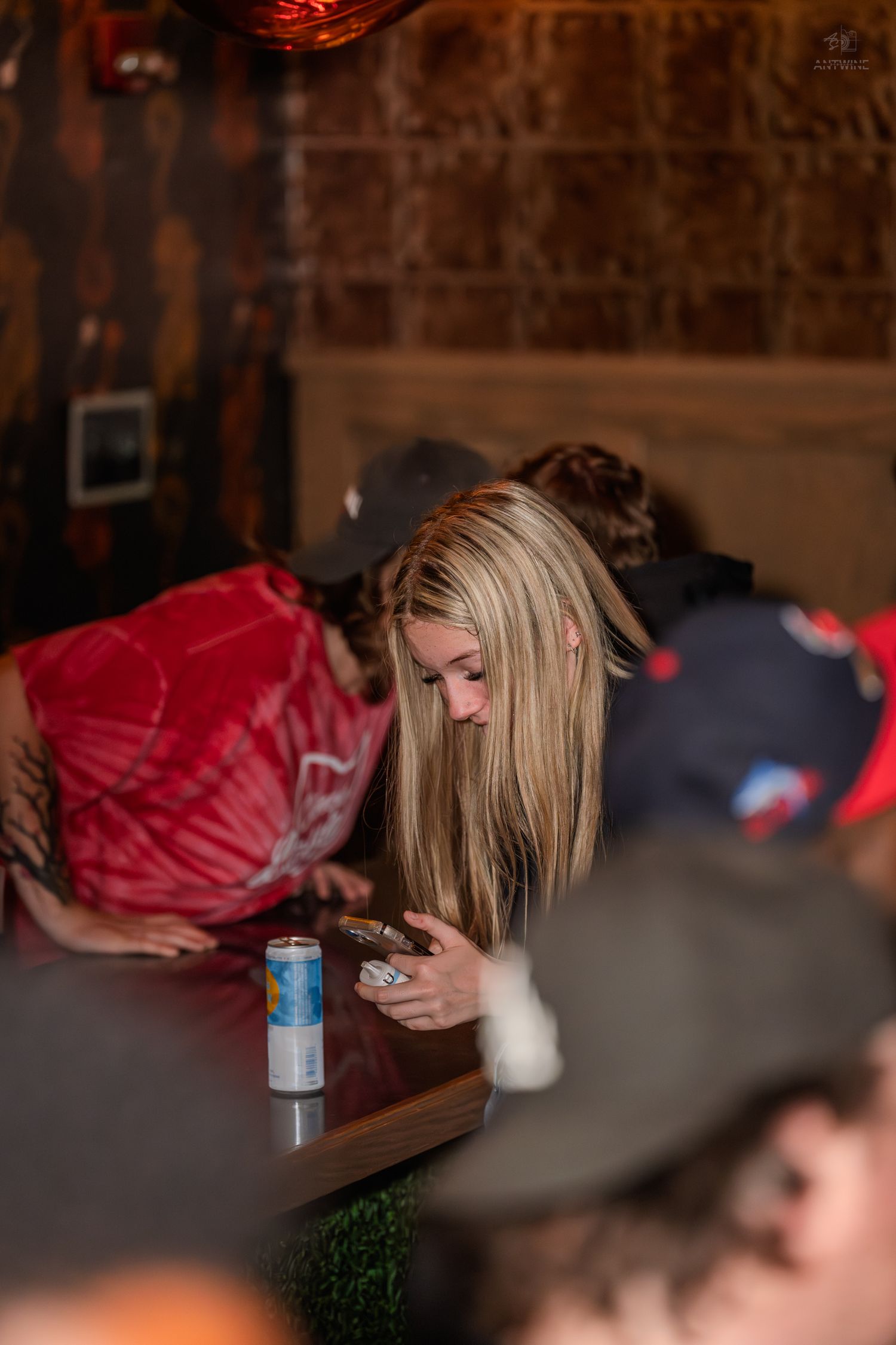 Blonde woman looking at her phone at a dimly lit bar with a Red Bull can nearby.