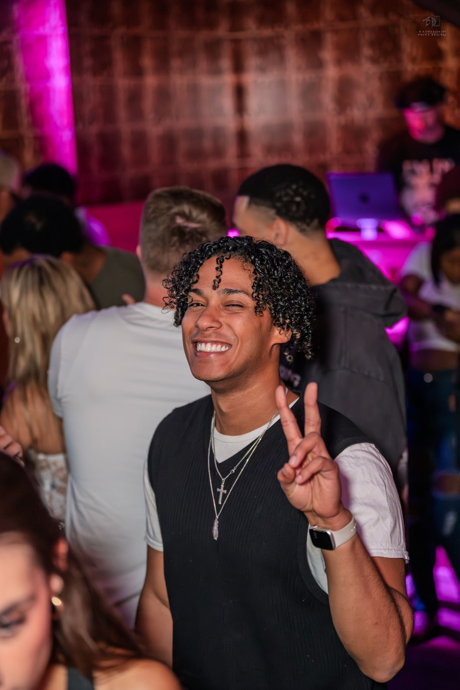 Young man with curly hair smiling and flashing a peace sign at a nightclub party.