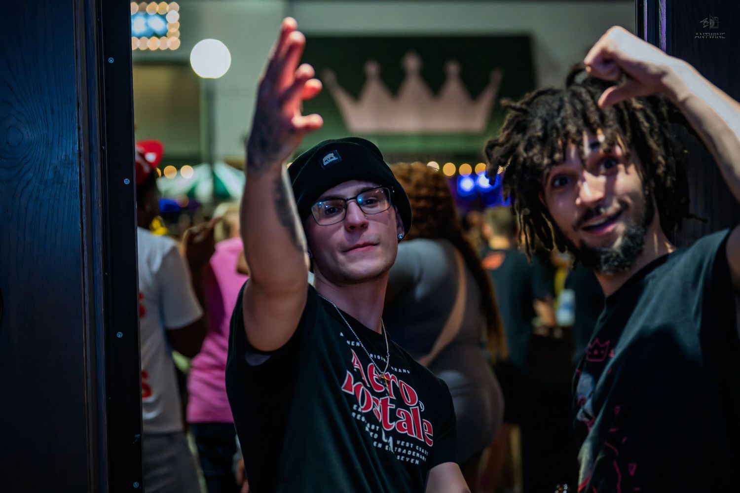 Two young men posing near a venue entrance with colorful lights behind them.
