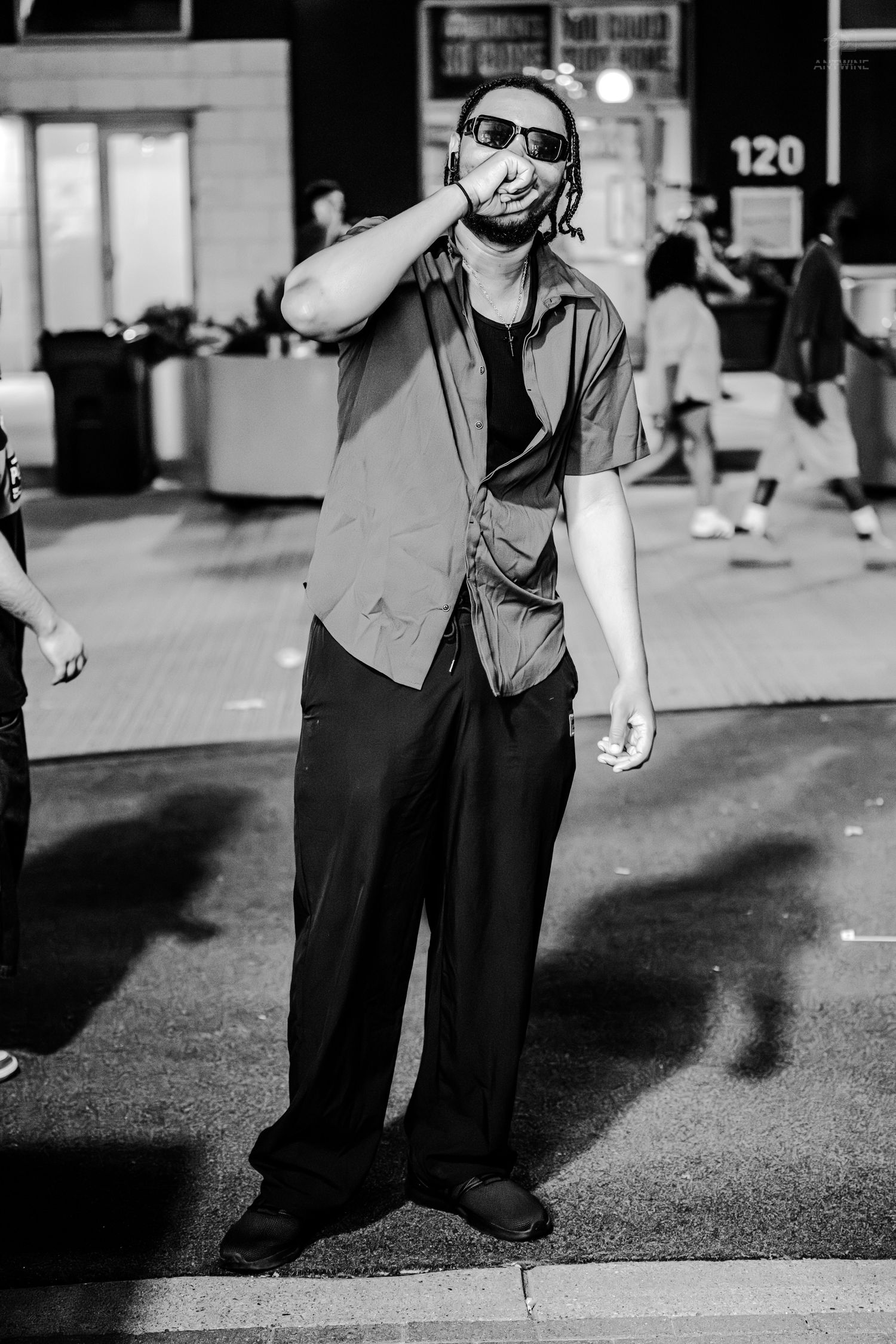 A man in sunglasses and a button-up shirt stands on a street at night in a black and white photo.