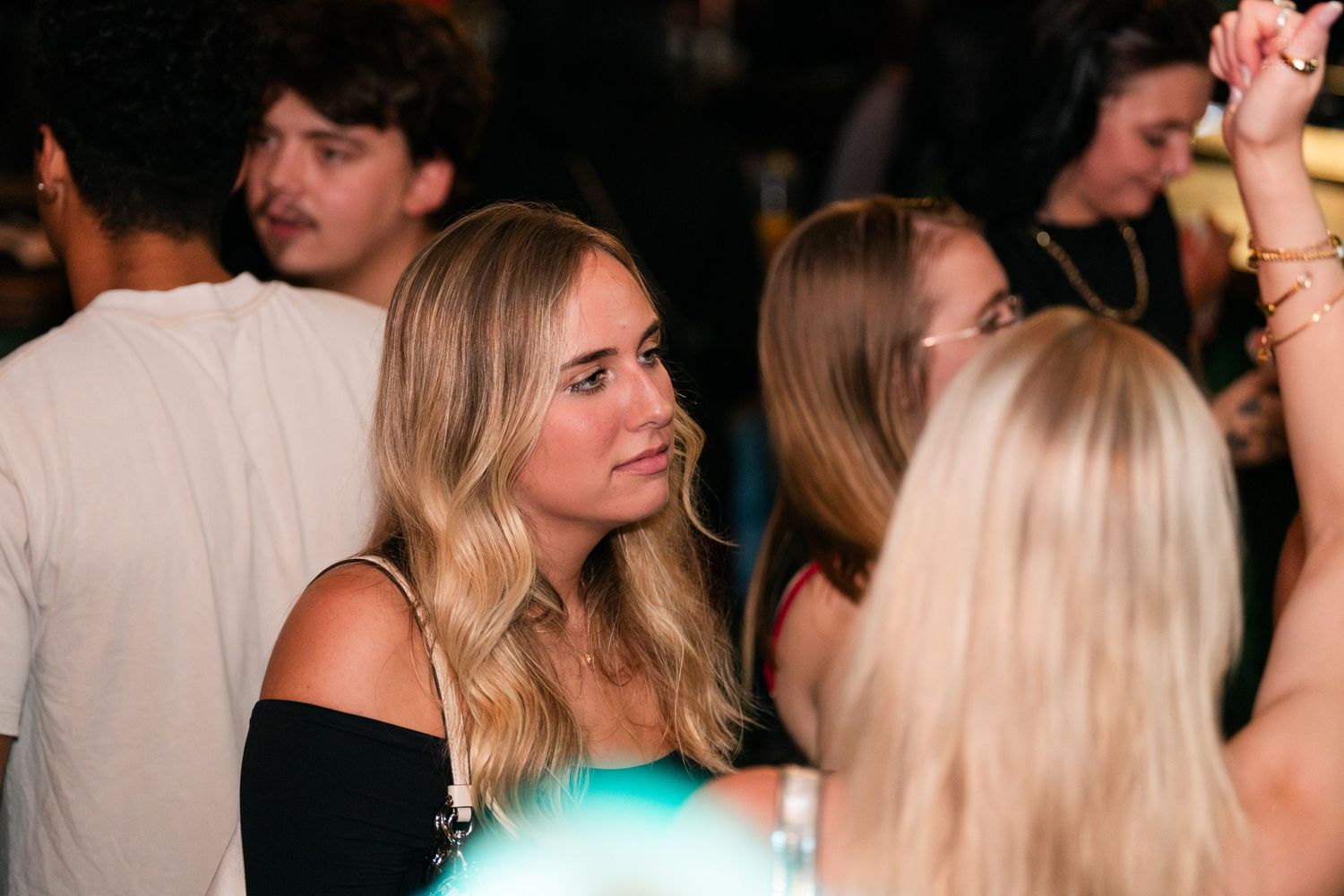 Young blonde woman in black top looking sideways at a dimly lit nightclub party.