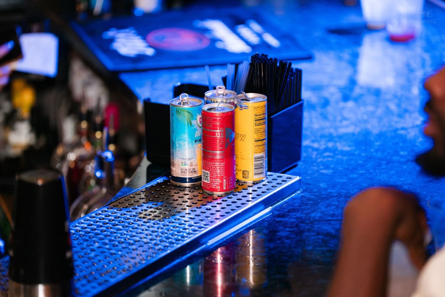 Colorful Red Bull cans displayed on a blue-lit bar counter with beer taps in background.