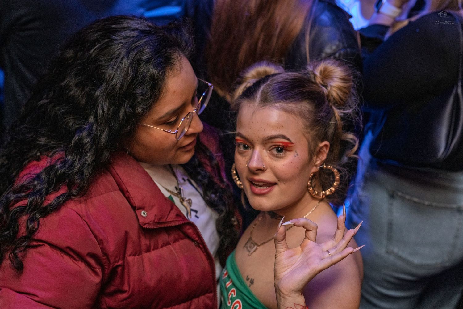 Two women smile and pose together at a nightclub, one wearing gold hoop earrings and face gems.