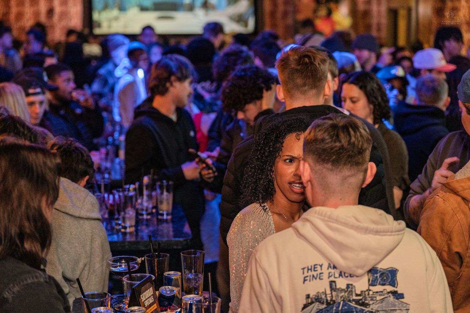 Diverse group of young adults socializing at a crowded bar with drinks on the table.