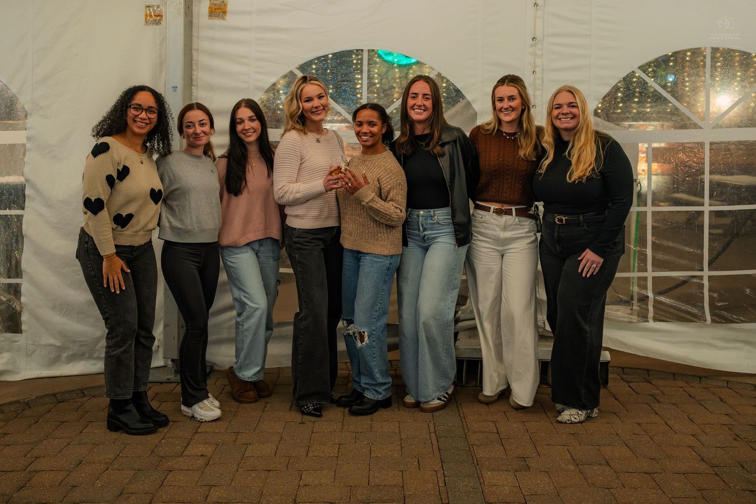 Group of eight young women smiling and posing together inside a white tent venue at a social event.