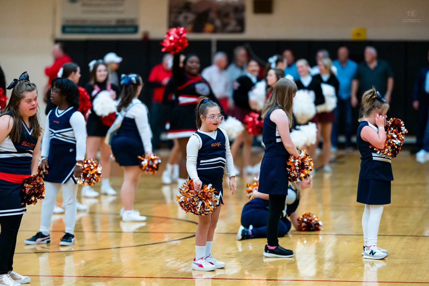 Young cheerleaders in navy uniforms smile and perform with gold pom-poms in a packed school gymnasium.