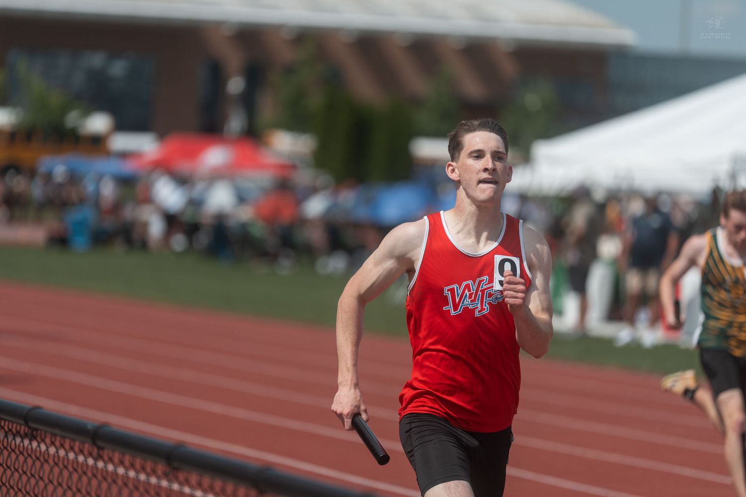 Male relay runner in red uniform sprints on outdoor track during competitive relay race event.