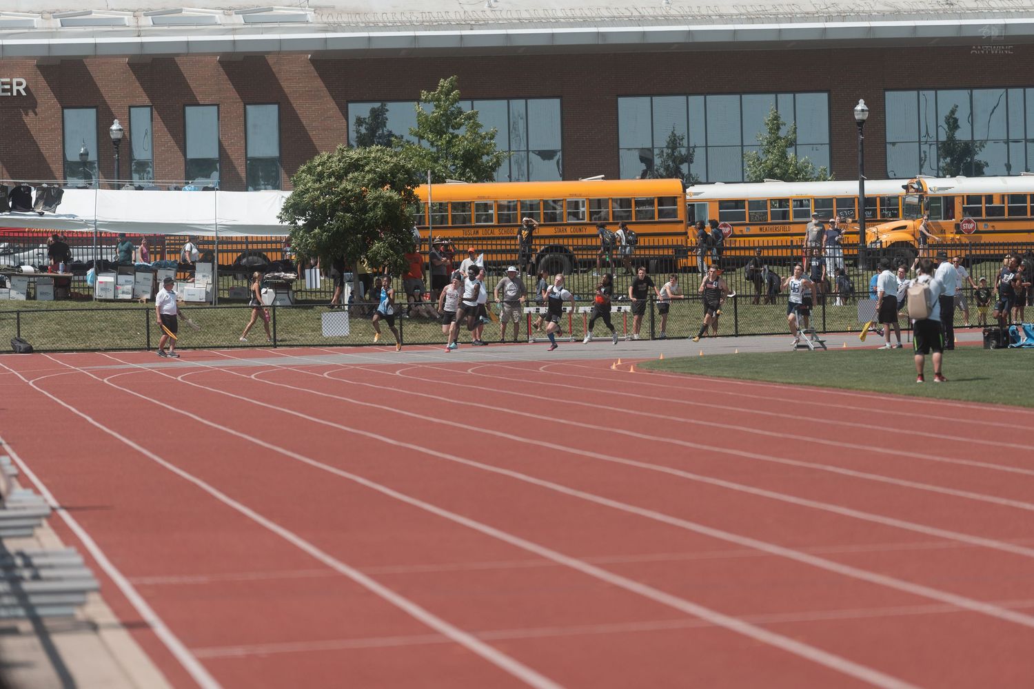 Athletes run on a red track during a high school track and field meet with school buses in background.