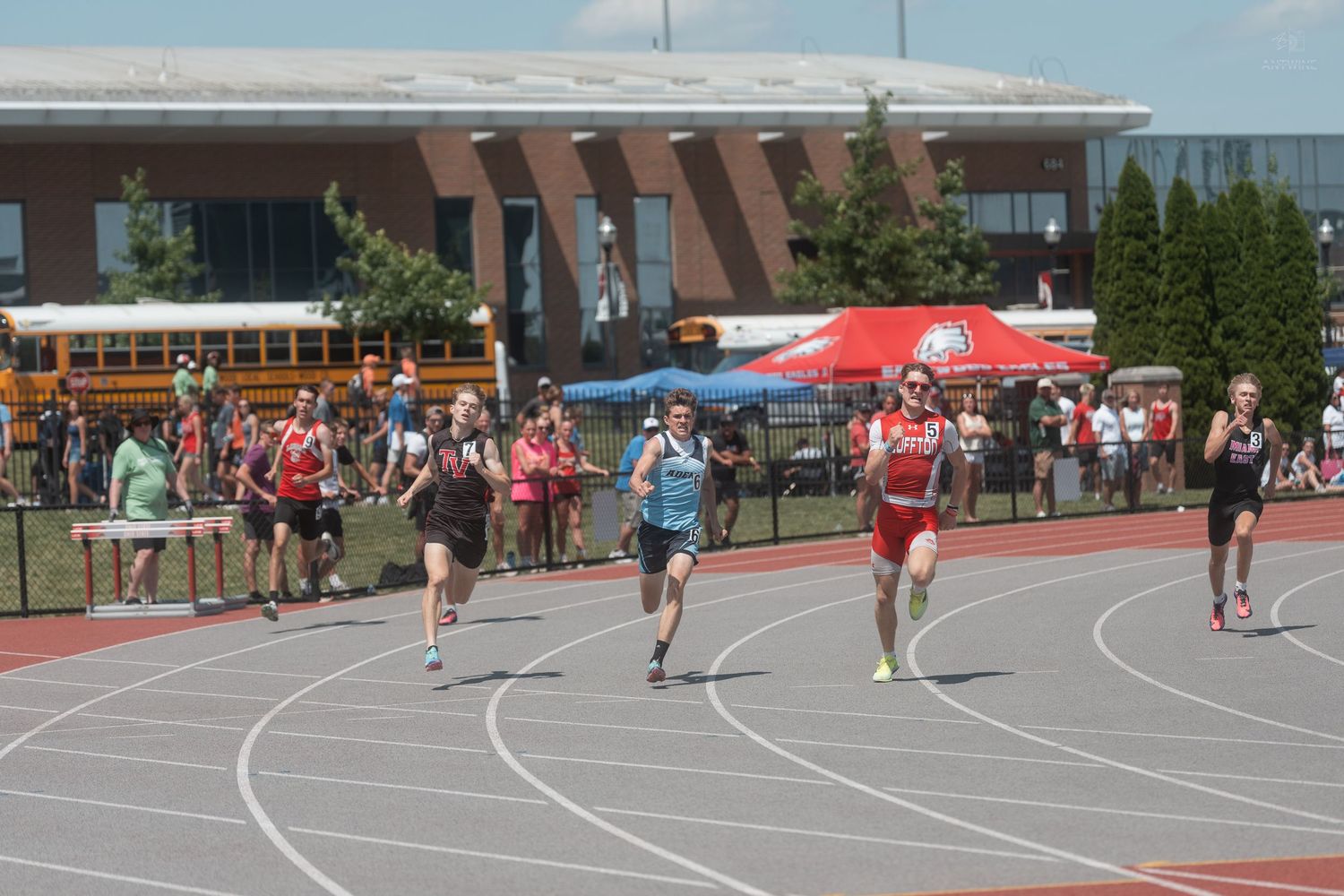 Athletes racing on a red track during a sunny outdoor high school track and field competition.