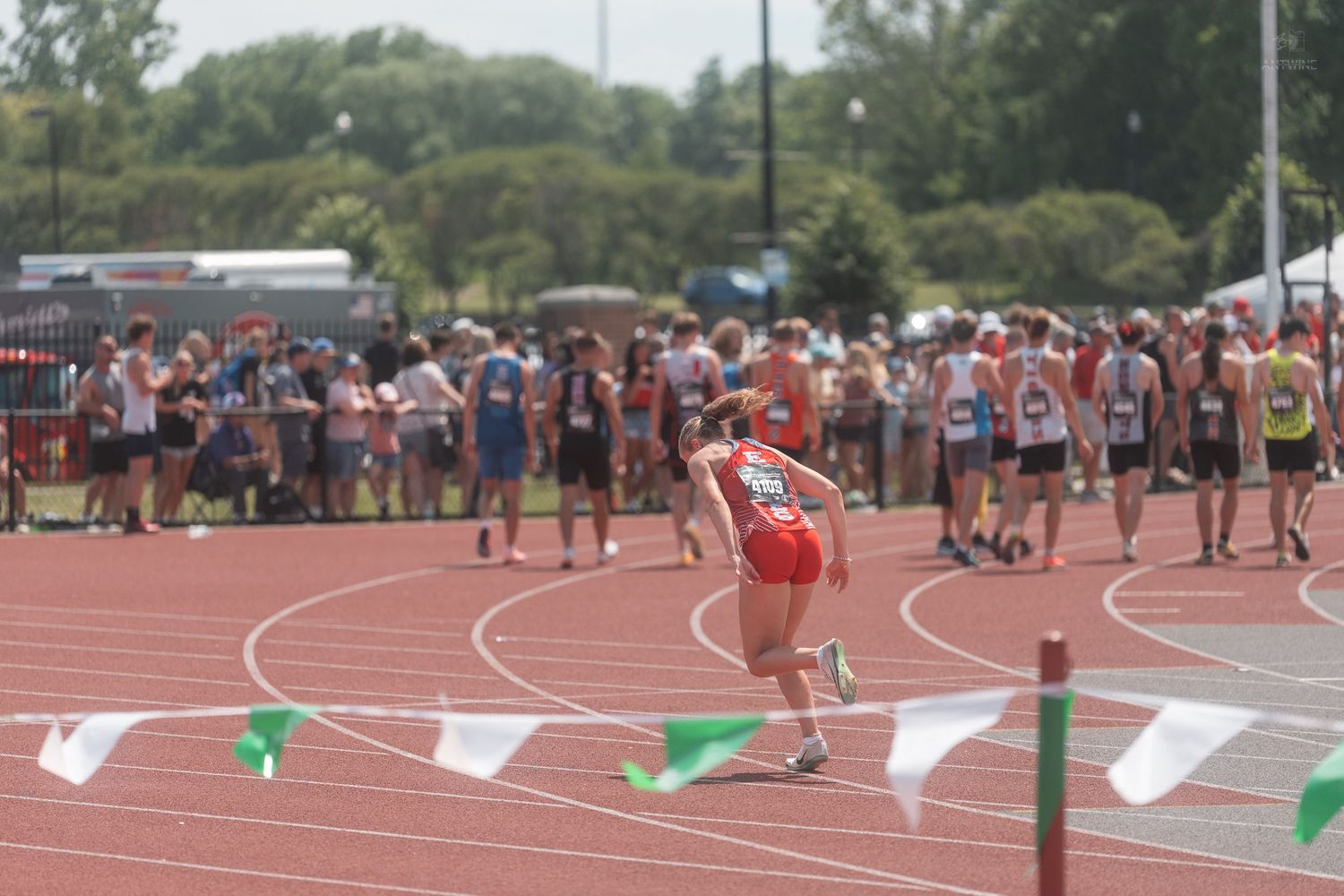 Runner in red uniform rounds track bend at large outdoor high school track and field meet.