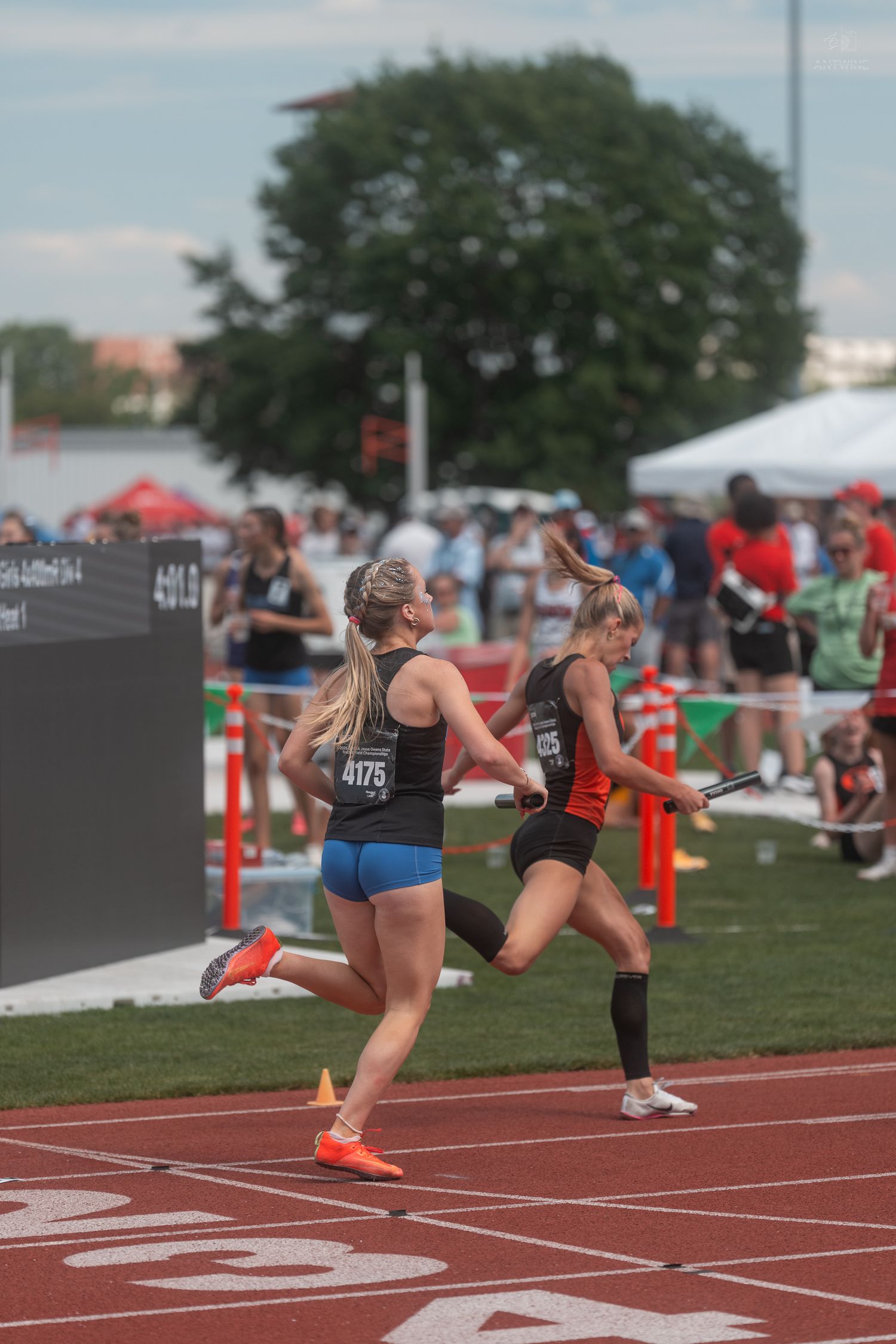 Two female track runners exchange a relay baton on a red track during a competitive high school track meet.