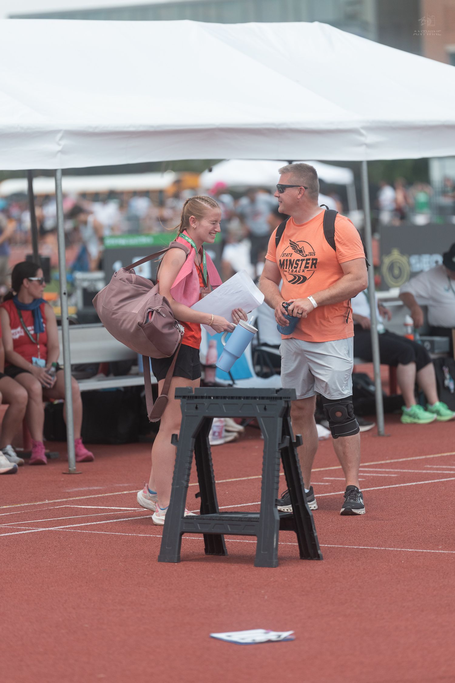A young female athlete and a coach in an orange shirt review documents at an outdoor track event.