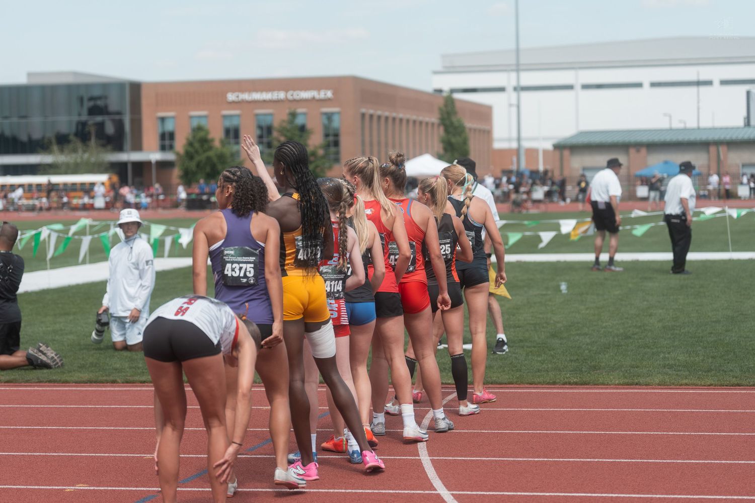 Female track and field athletes line up on the track before a relay race at a state championship meet.