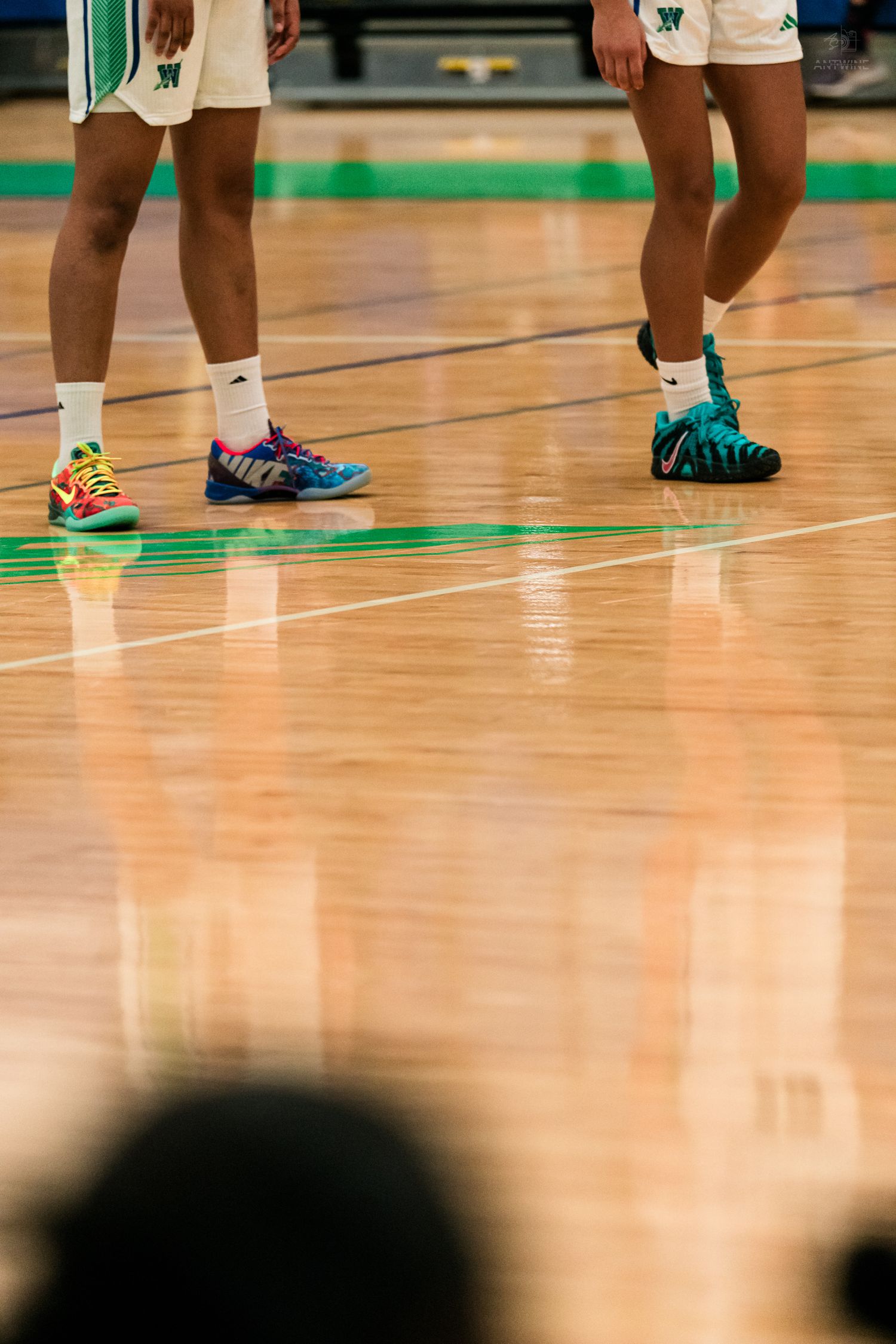 Two Warriors basketball players stand on polished hardwood court wearing colorful mismatched sneakers.