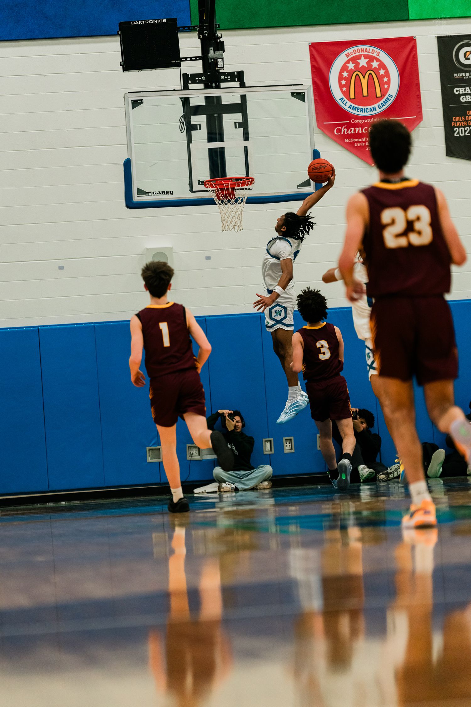 A Warriors player drives to the basket for a layup against maroon-uniformed defenders in a packed gymnasium.