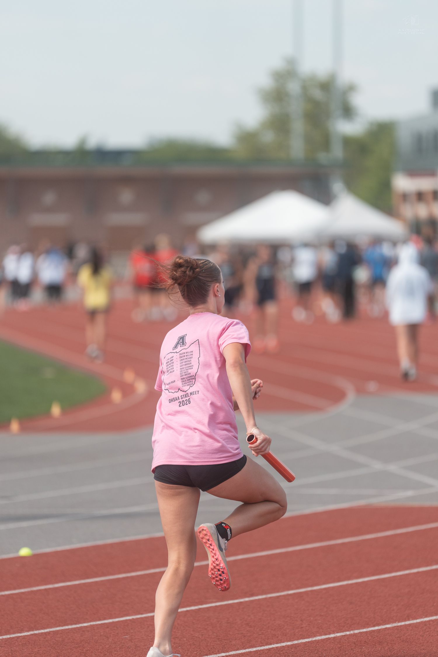 Female athlete in pink shirt runs relay race with baton on red outdoor track.