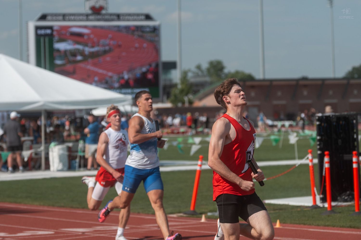Exhausted runner in red crosses the finish line at an outdoor high school track and field championship.