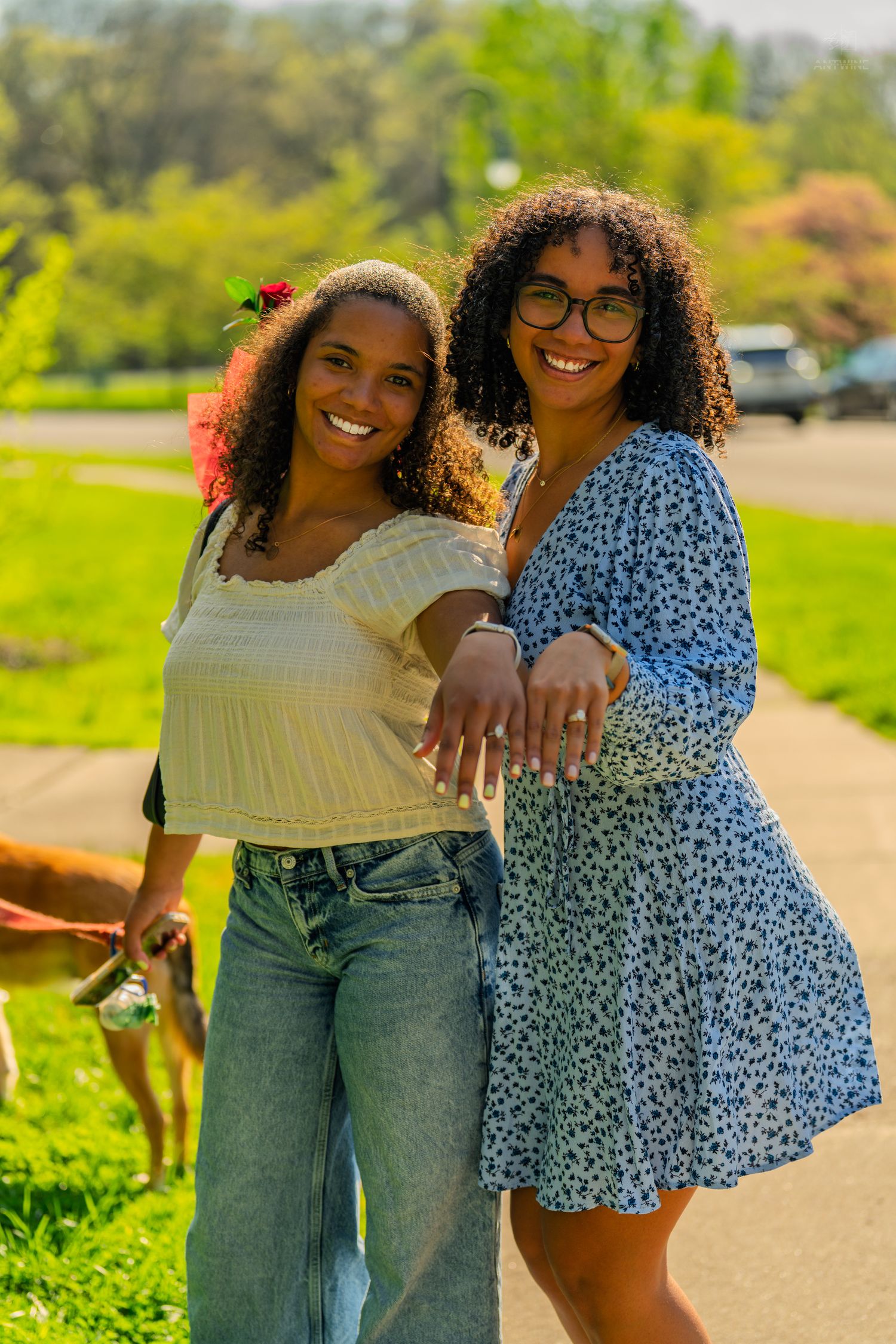 Two smiling women with curly hair pose outdoors in a sunny park, one in jeans and crop top, other in blue floral dress.