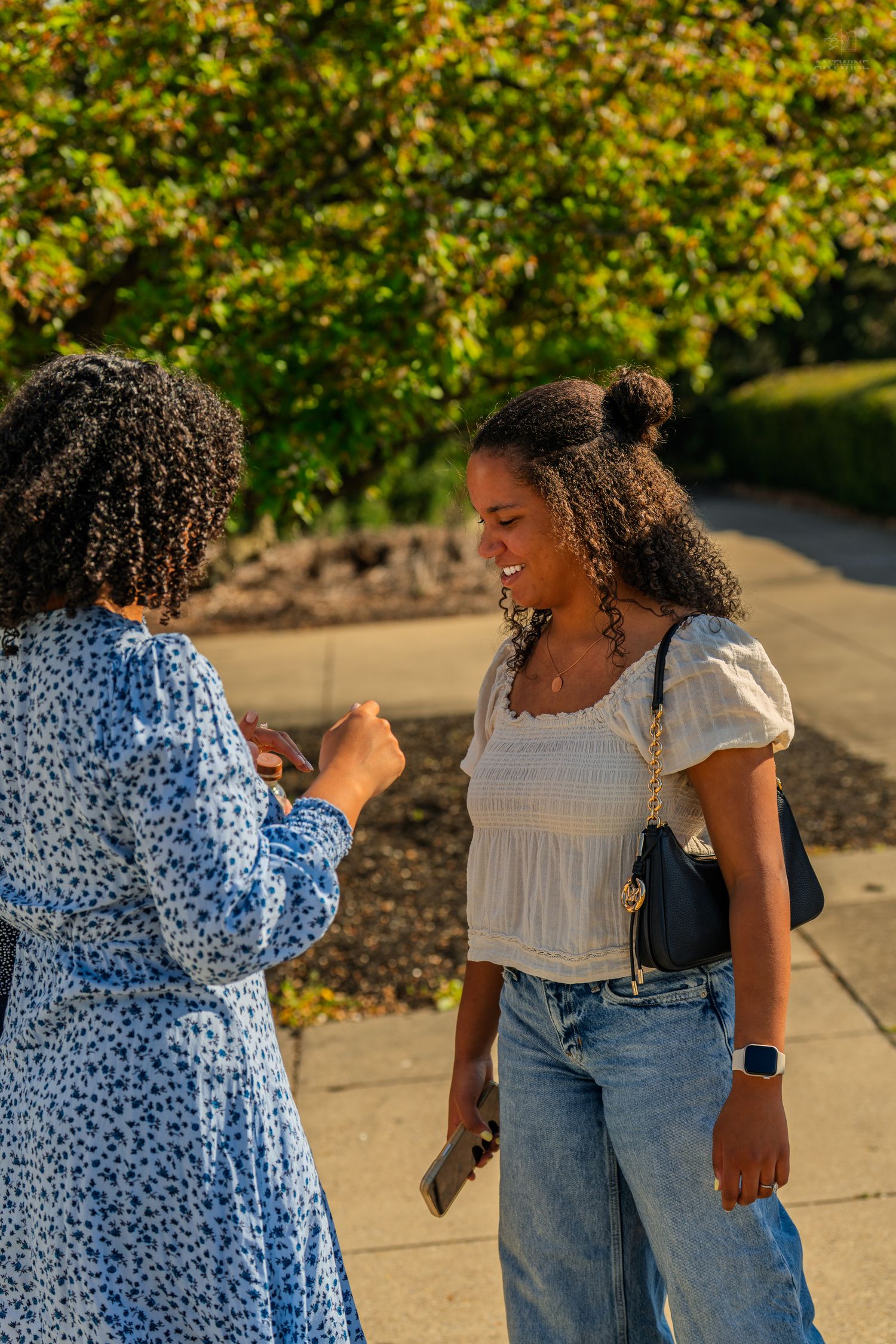 Two young Black women smiling and talking outdoors near colorful autumn trees.