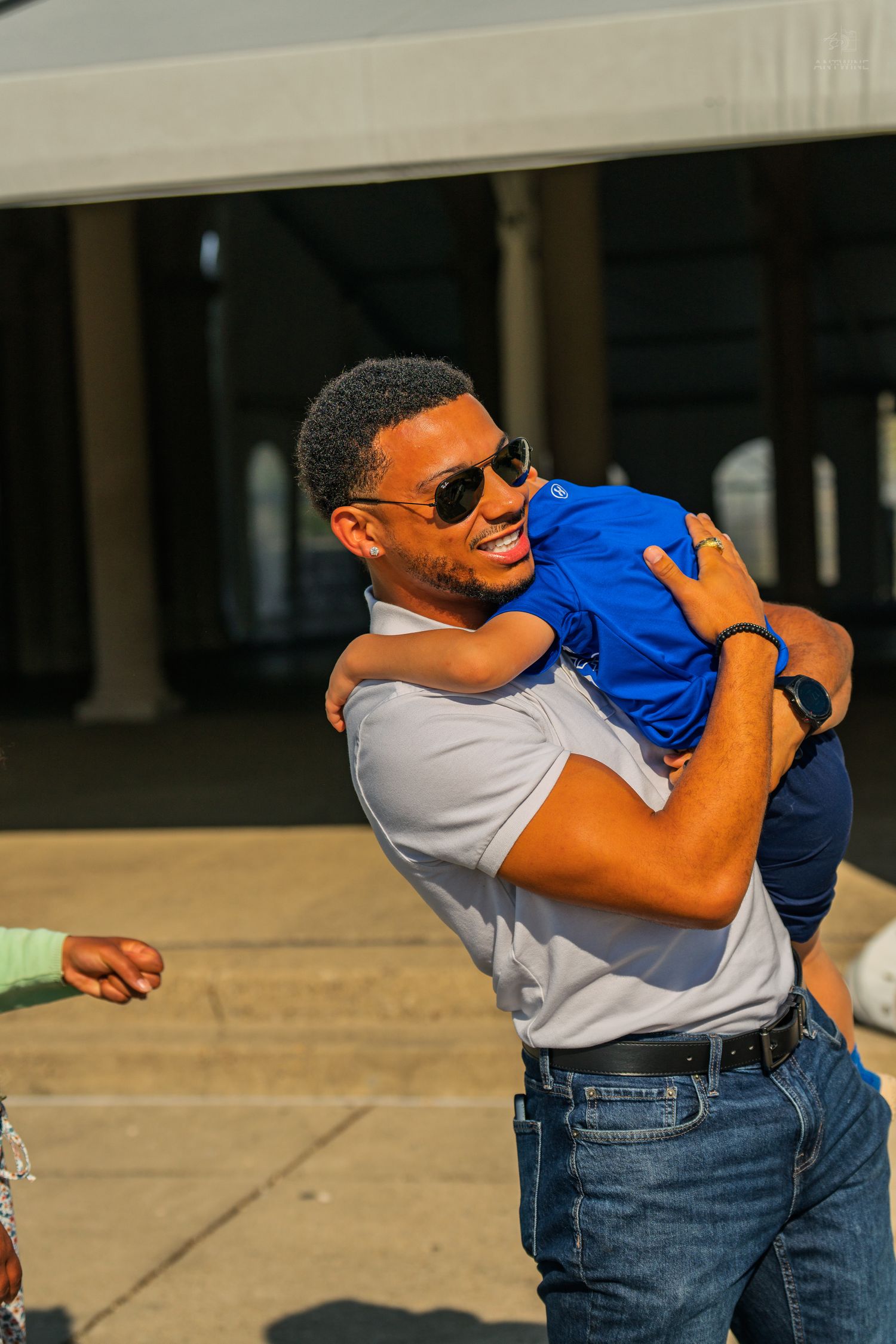 A laughing man in sunglasses joyfully carries a young child in a blue shirt outdoors in bright sunlight.