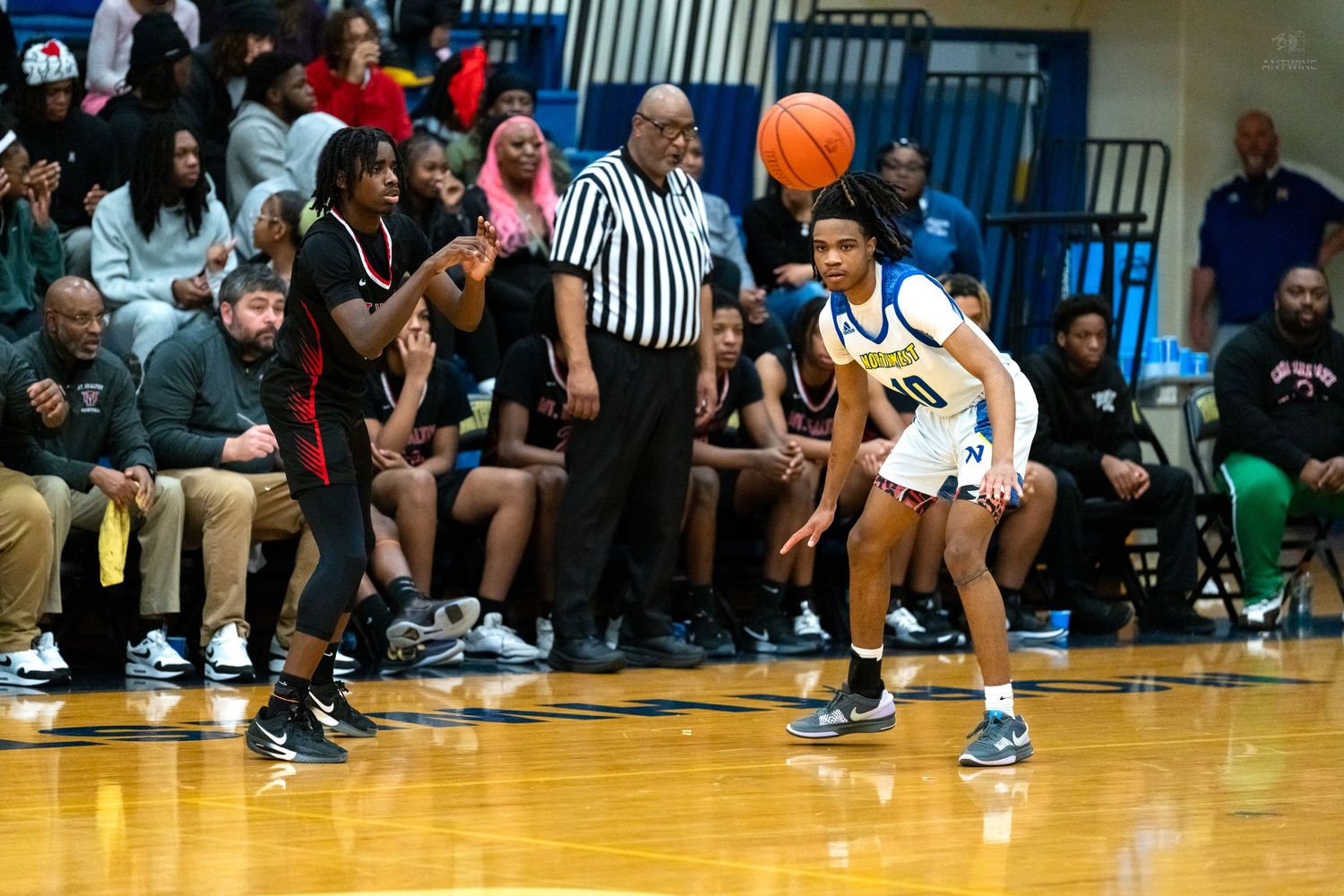 Young basketball player in white uniform dribbles past defender with referee and crowded bleachers in background.