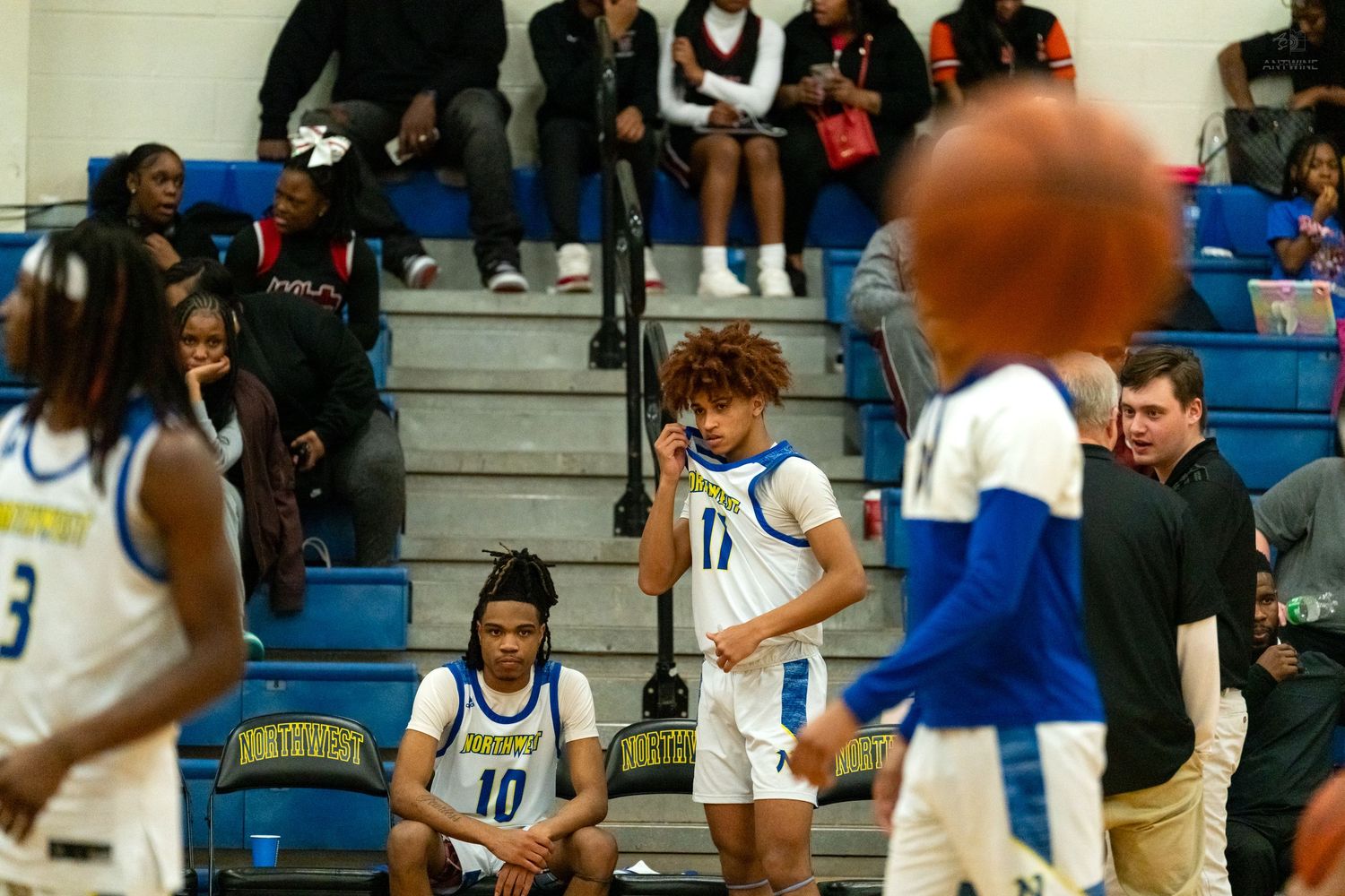 High school basketball players in blue uniforms rest on bench while a coach stands nearby during a timeout.