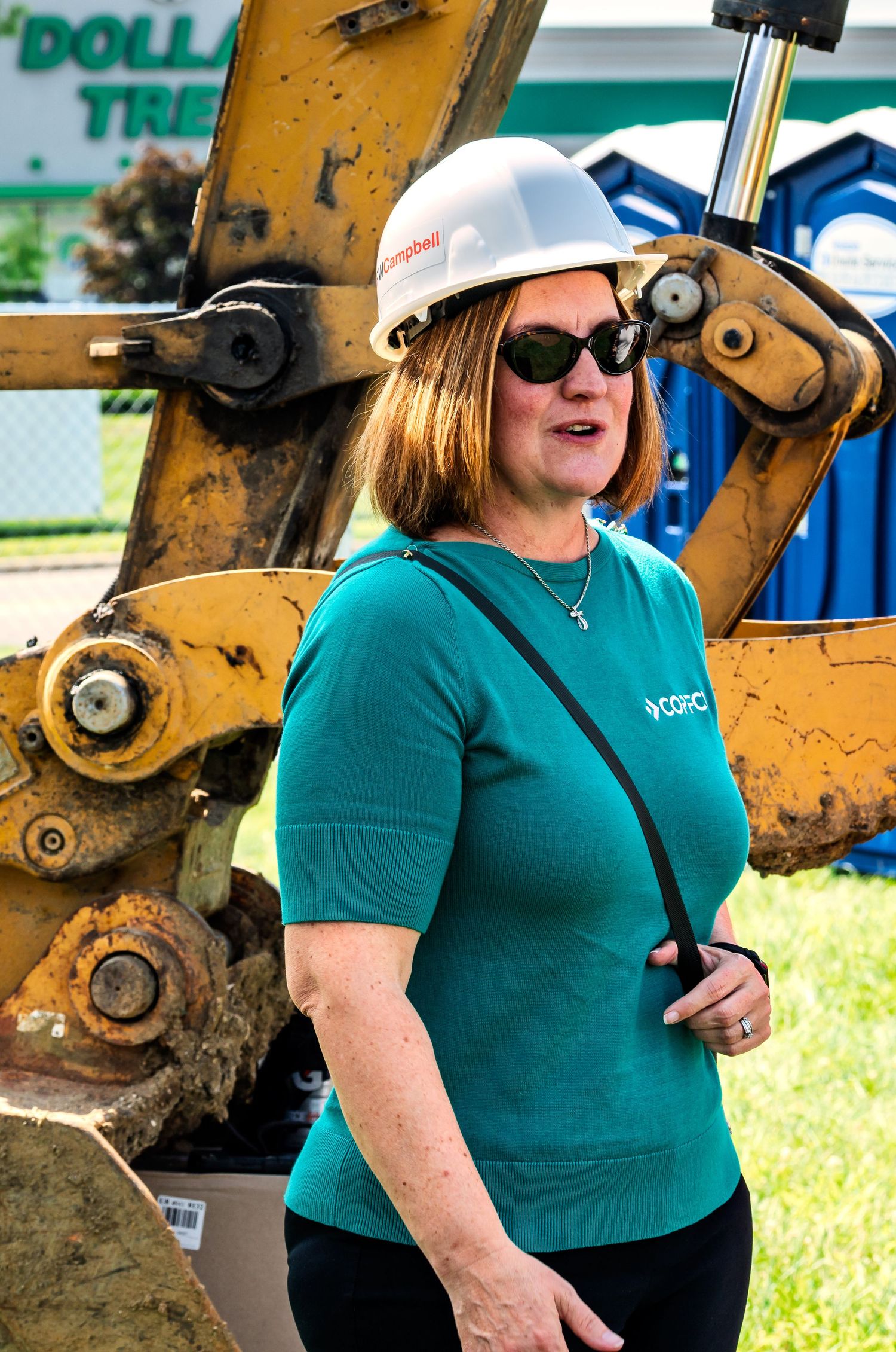 Woman in teal COPFCU shirt and hard hat stands near yellow construction excavator at job site.