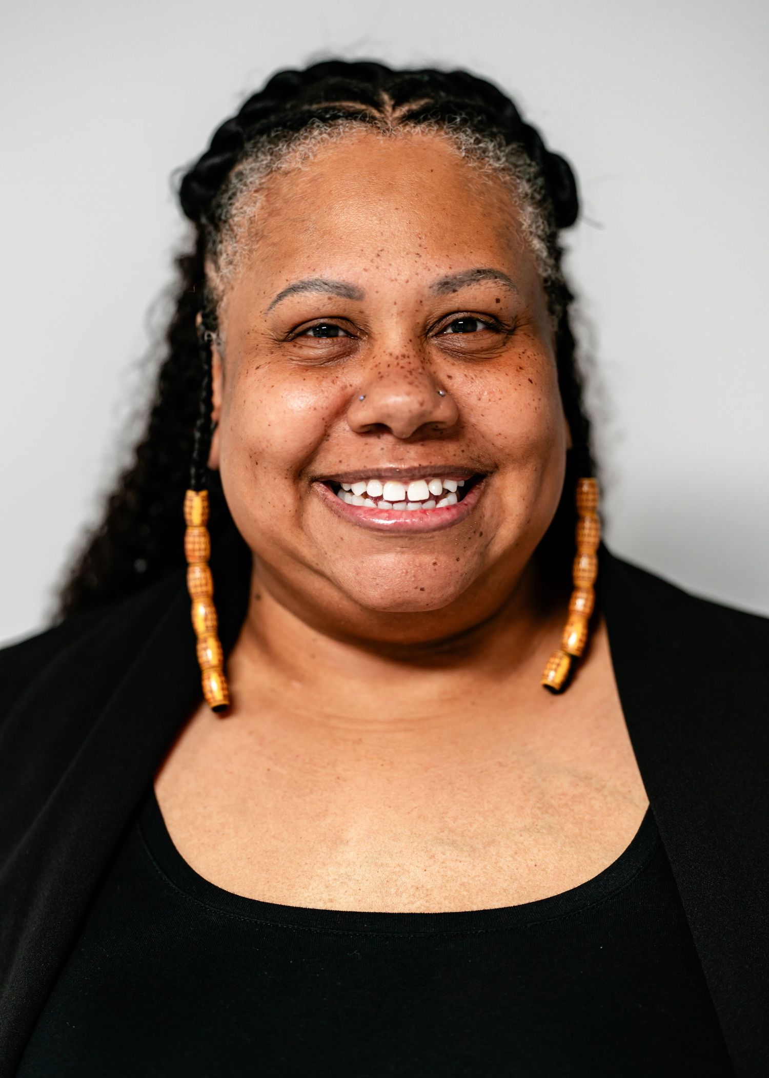 Joyful woman with braided hair and gold hoop earrings smiling broadly in a black outfit.