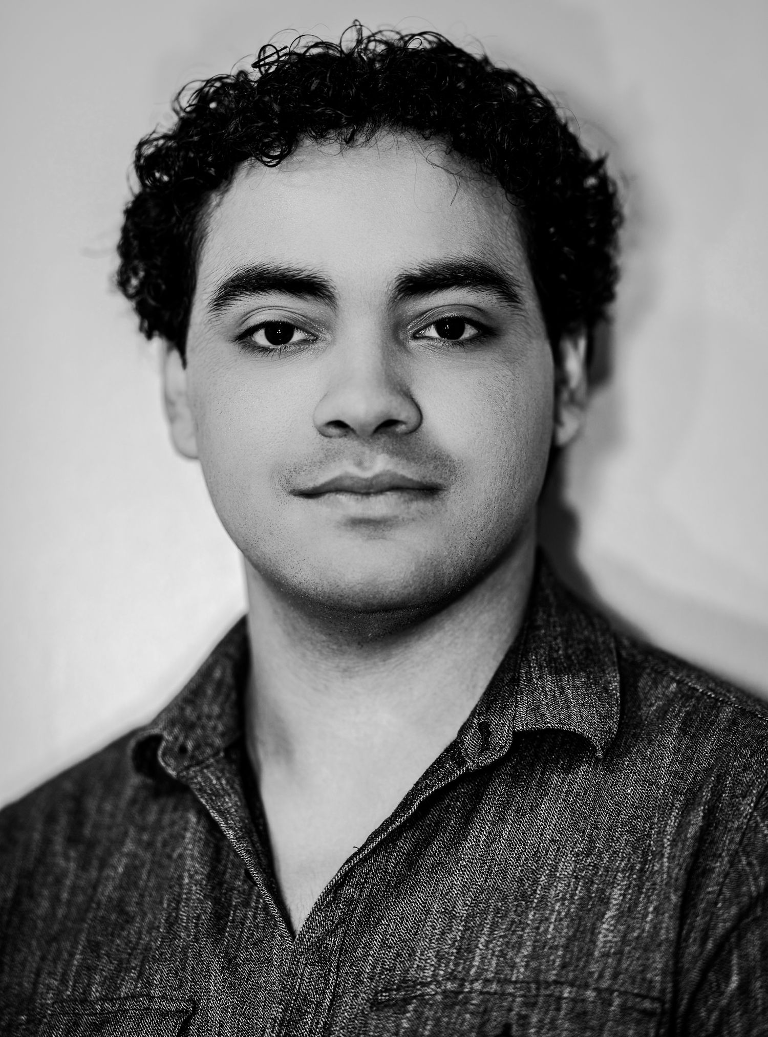 Young man with curly dark hair in a dark shirt looking serious in a black and white portrait.